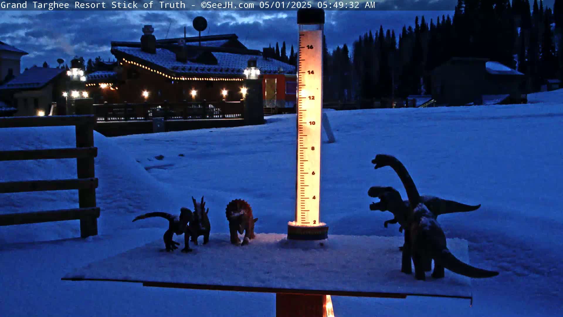 Several dinosaur figurines are arranged on a snow-covered surface in front of a lit building at twilight, next to a tall illuminated rain gauge.