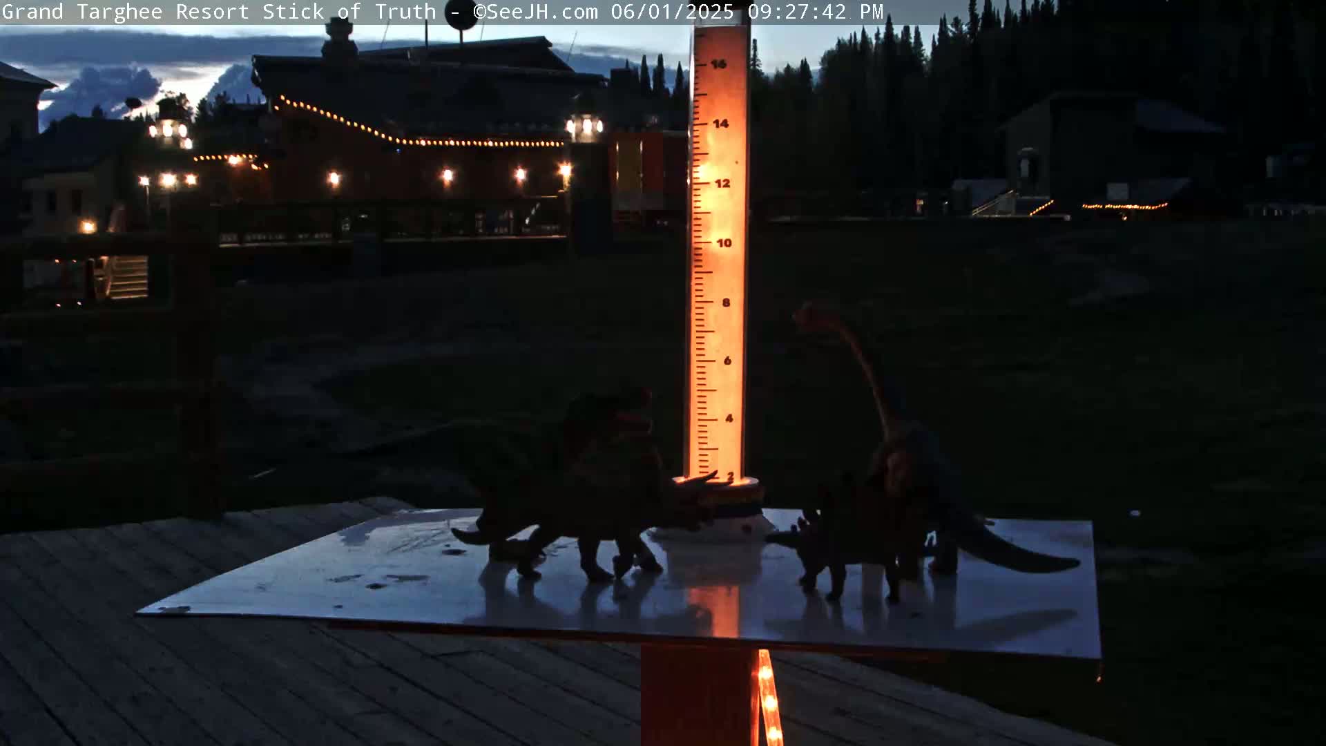 At night, several dinosaur toys are arranged around a tall, illuminated measuring stick on a deck, with a dark building and trees in the background.