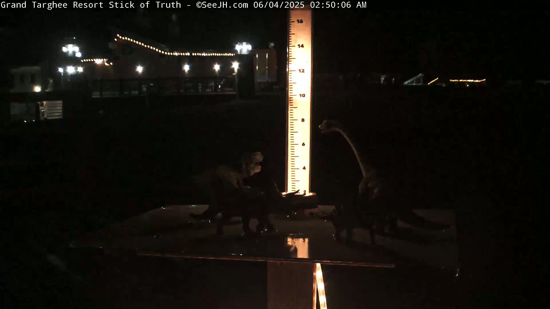At night, three dinosaur figurines are positioned near a tall illuminated measuring stick in front of a dimly lit building.