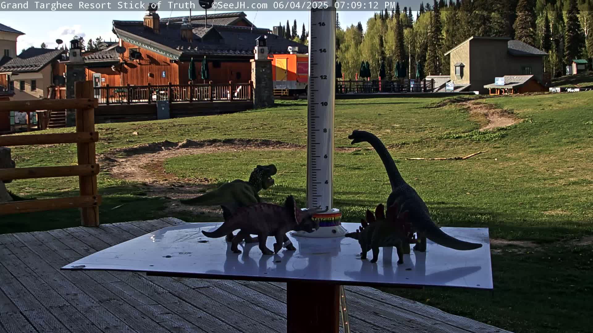 Several plastic dinosaur toys are arranged around a weather instrument on a deck overlooking a grassy area with buildings in the background, under a partly sunny sky.