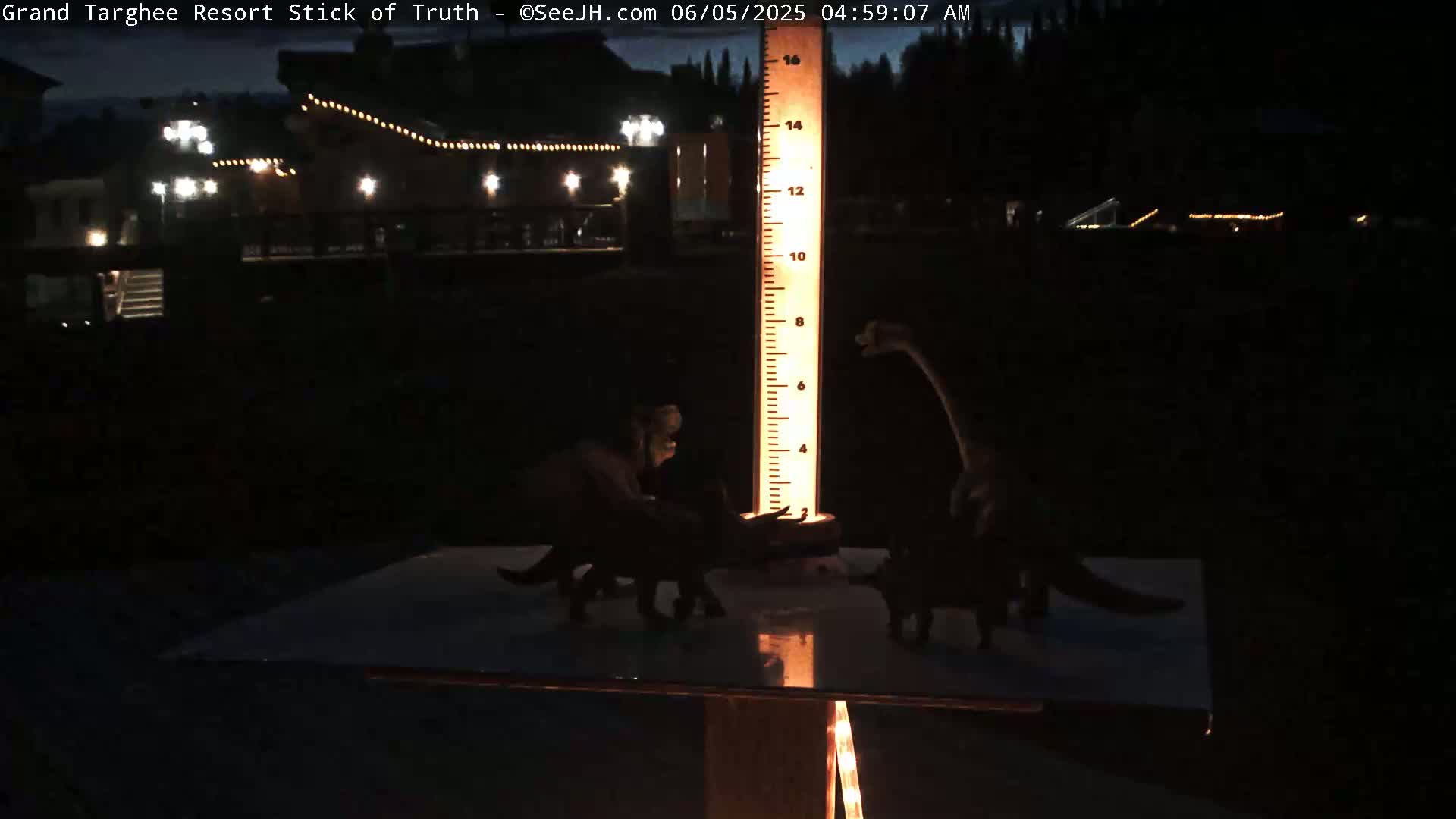 At night, illuminated toy dinosaurs are arranged around a tall, illuminated measuring stick in front of a dark building with lights.