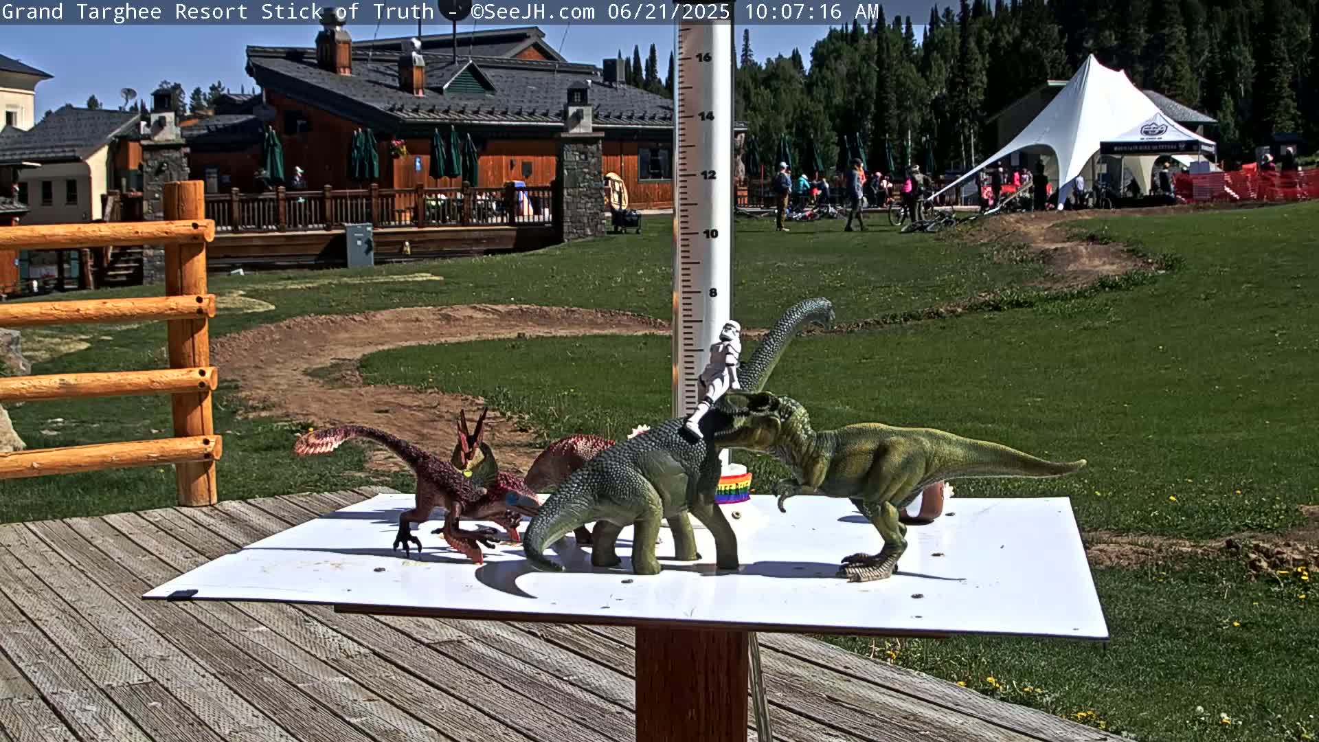 Several toy dinosaurs and a stormtrooper figurine are arranged on a white square surface attached to a wooden post in front of a wooden building and grassy area on a sunny day.