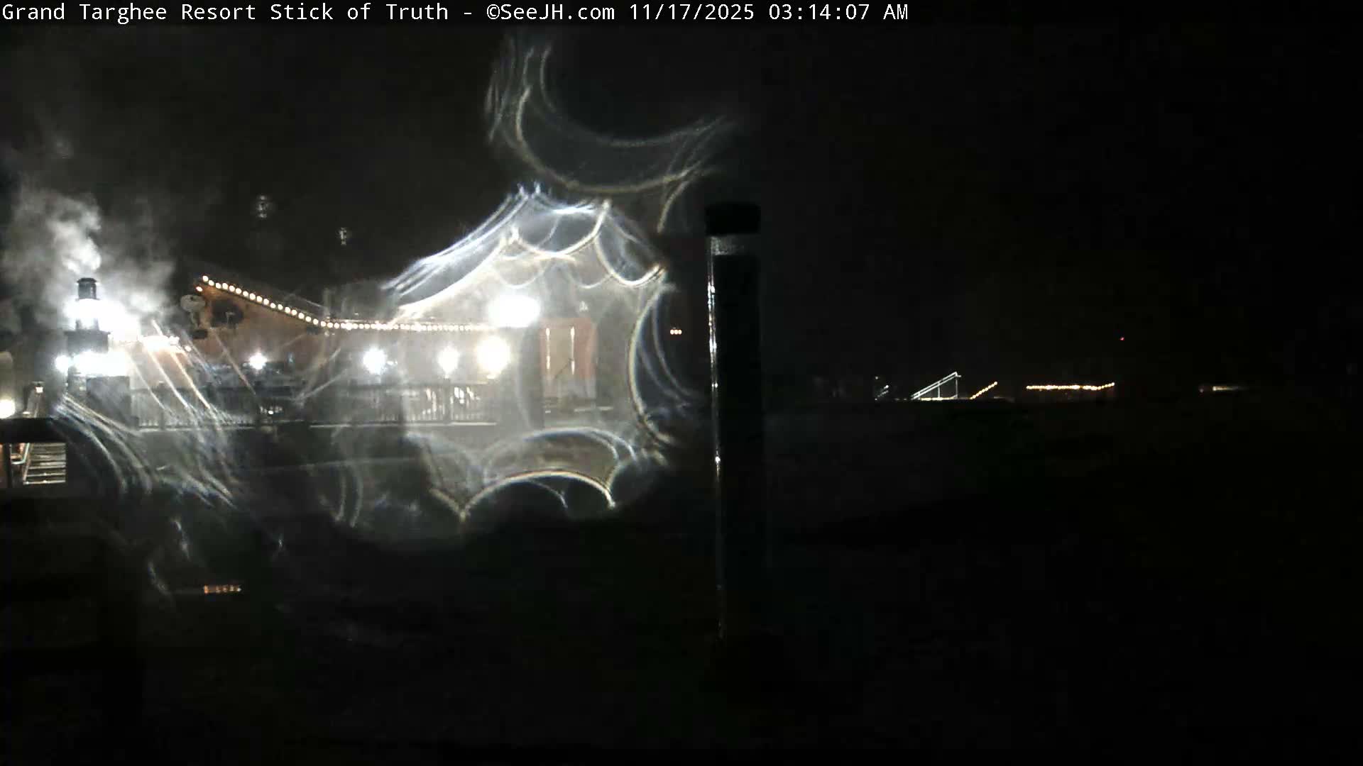 On a dark and humid night, a brightly lit resort lodge with steam rising from a chimney is partially obscured by prominent moisture streaks on the camera lens.