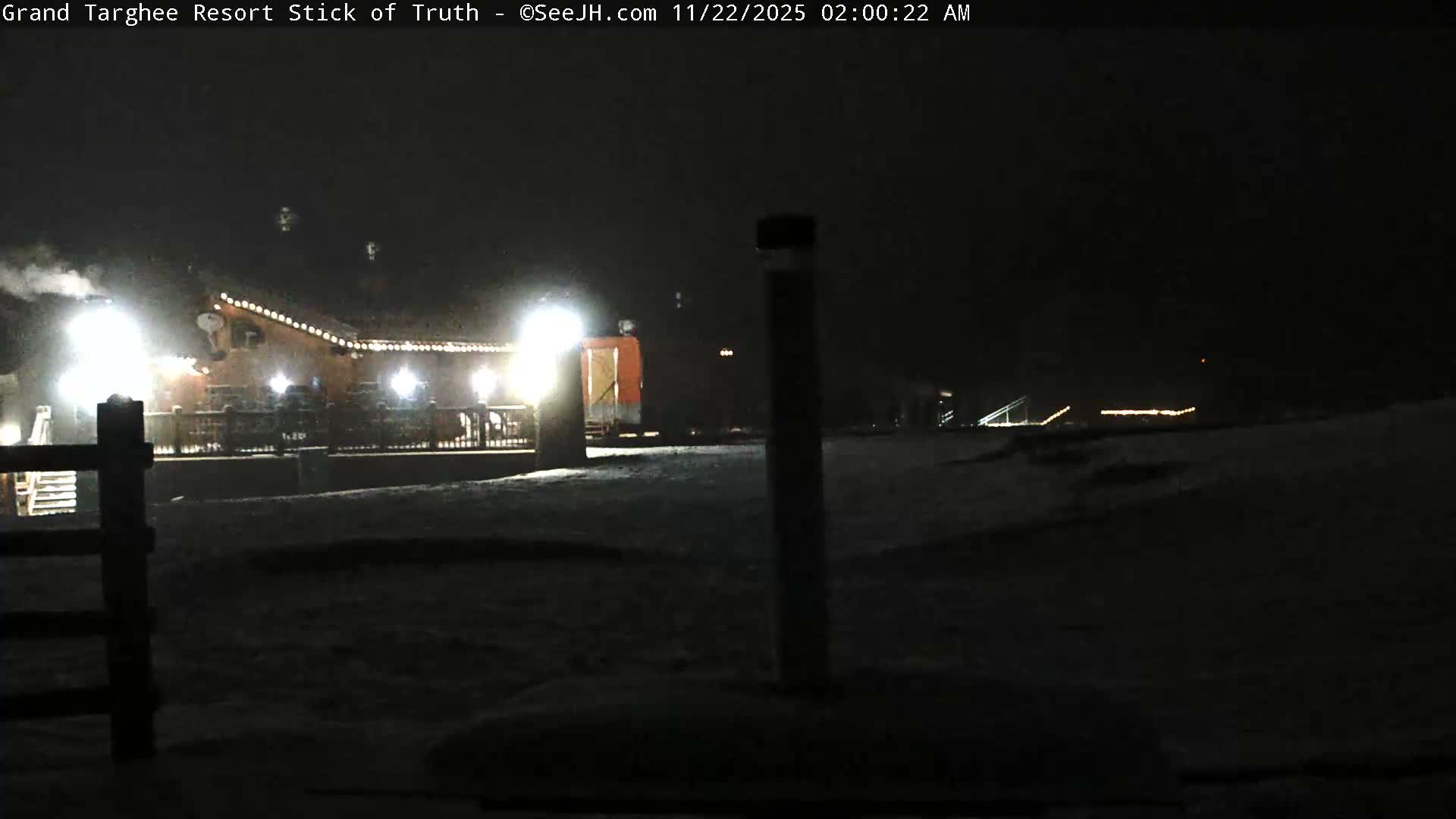 A brightly lit resort building adorned with string lights stands on a snow-covered landscape under a dark, clear night sky, with steam visible rising from a chimney.