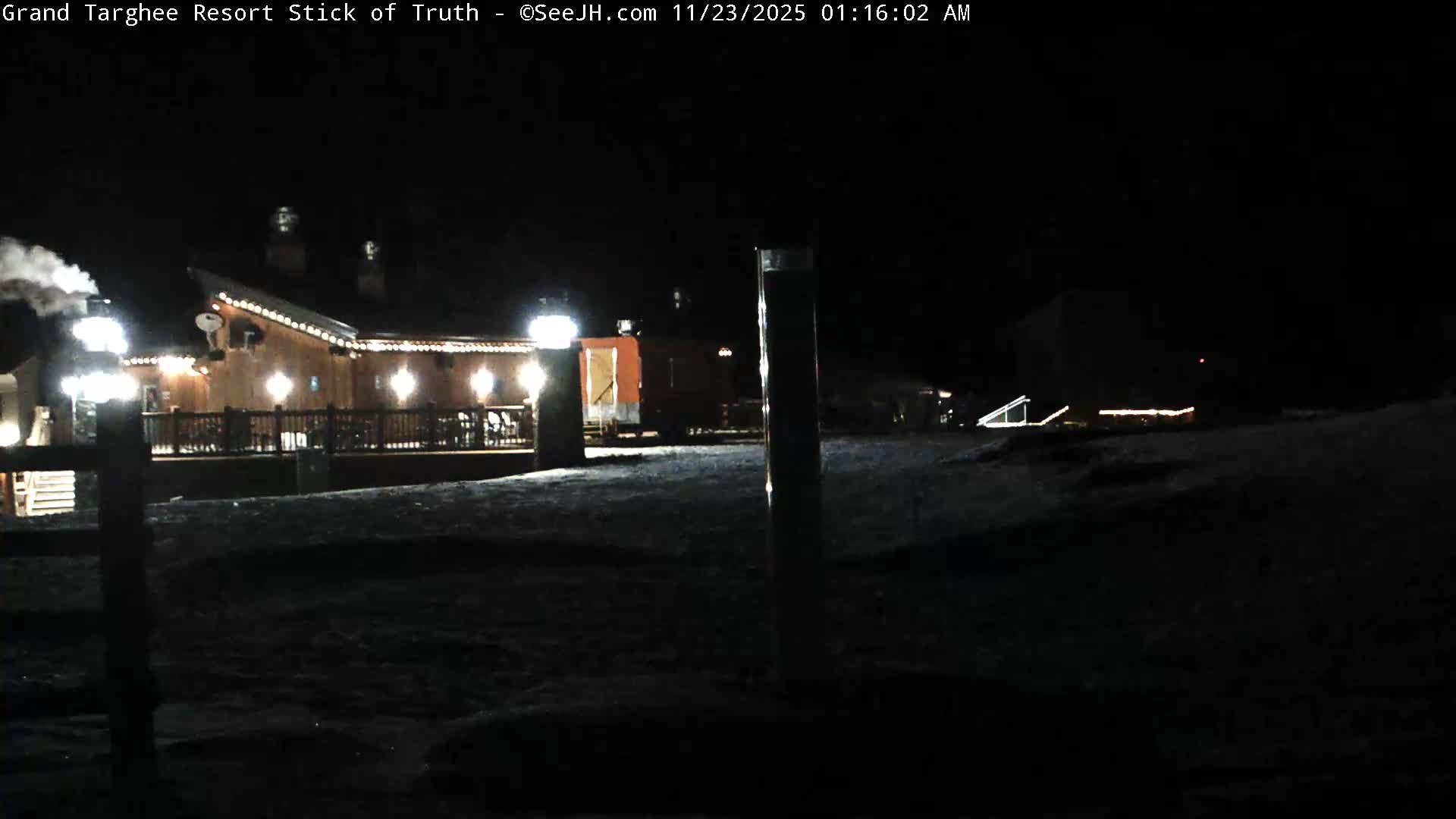 A well-lit wooden lodge with steam rising from its chimney stands on snow-covered ground under a clear, dark night sky.