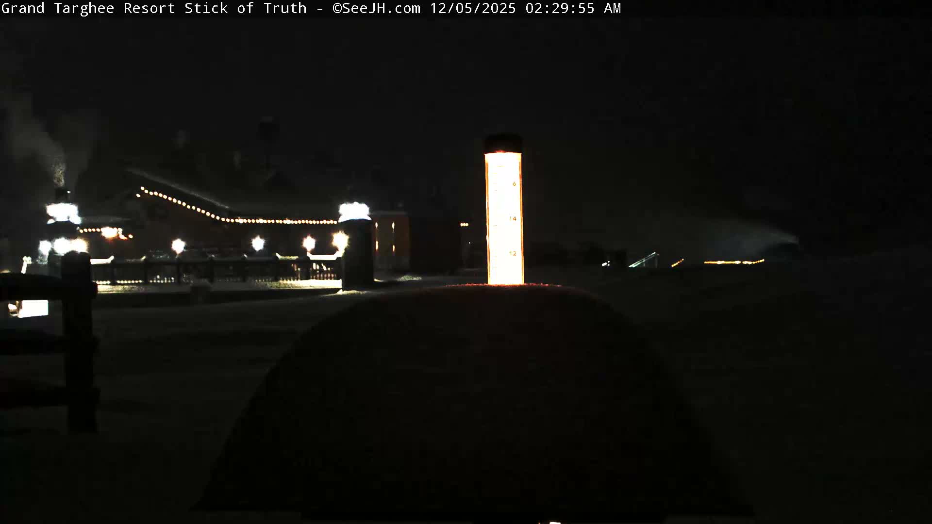 A cold, dark winter night at a snow-covered resort features an illuminated snow depth marker in the foreground, with distant buildings adorned with lights and smoke rising from a chimney.