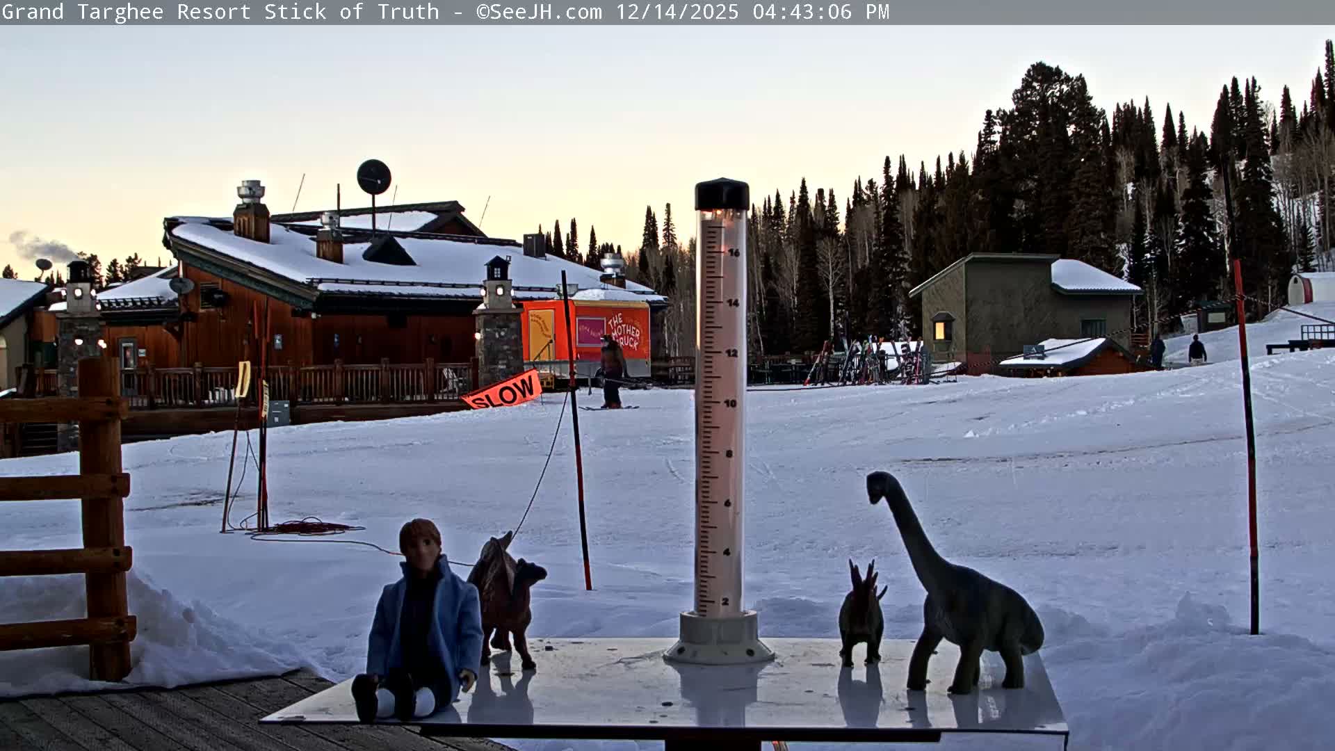 A cold, dark winter night at a snow-covered resort features an illuminated snow depth marker in the foreground, with distant buildings adorned with lights and smoke rising from a chimney.