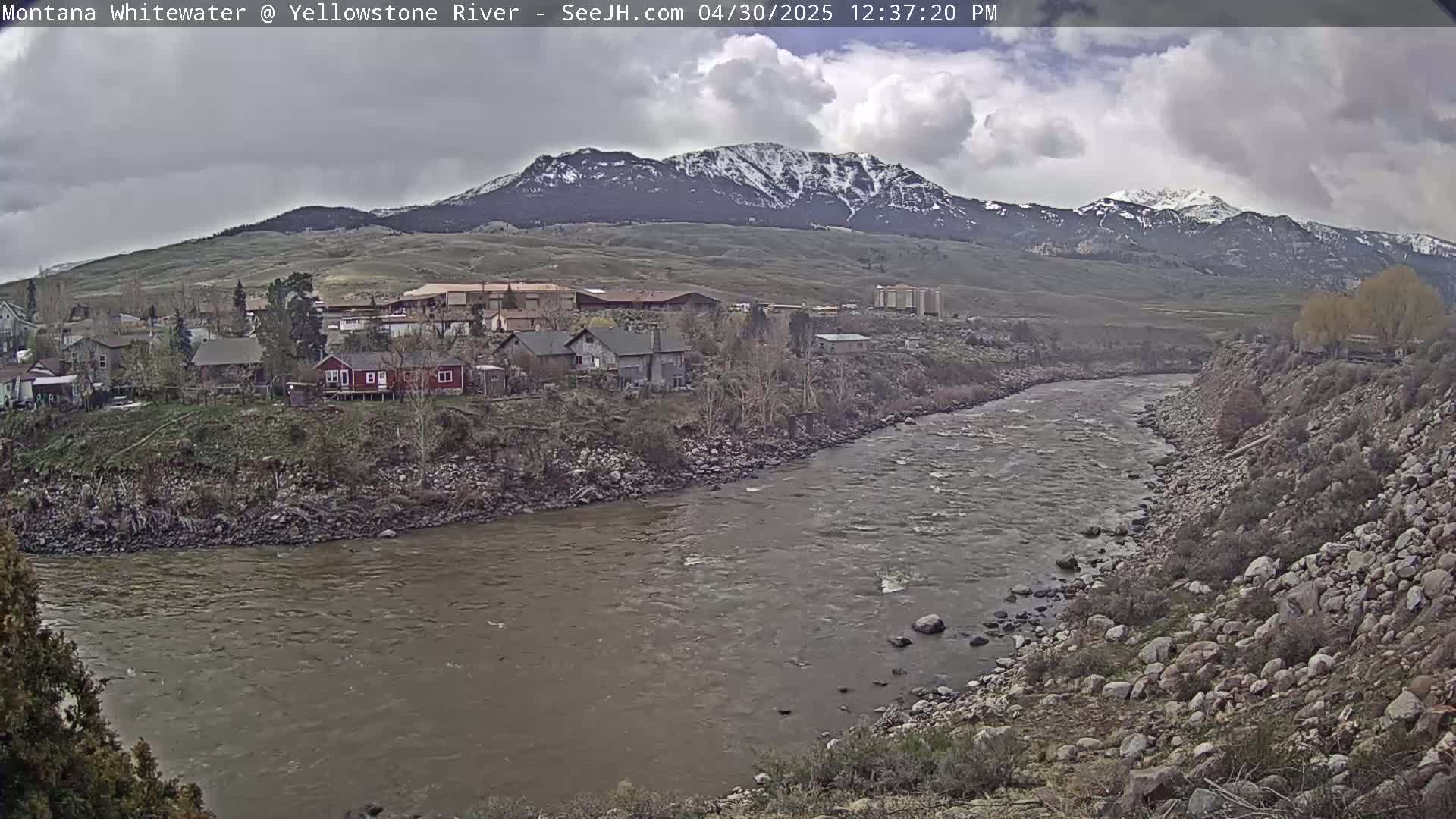 A cloudy day shows a wide, muddy river curving past a small town nestled beneath snow-capped mountains.