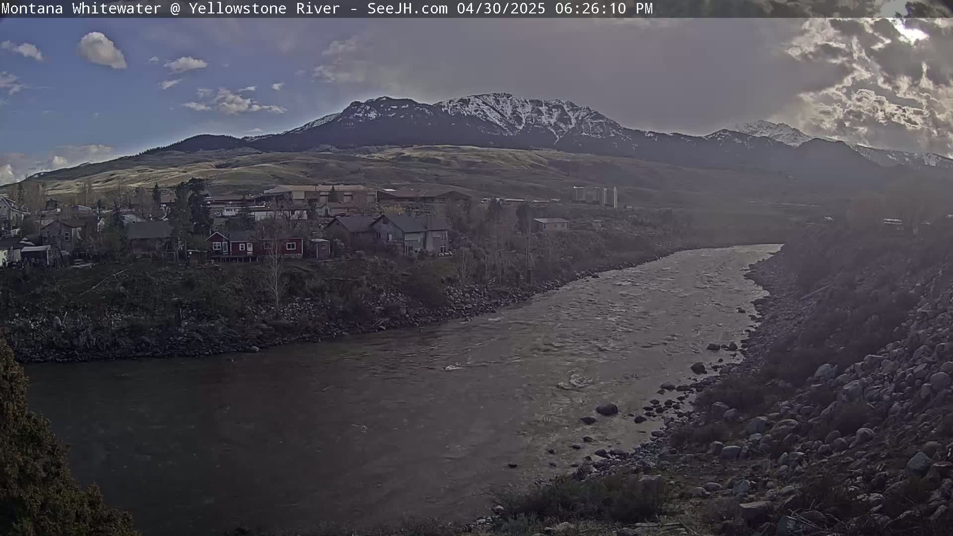 A river flows past a small town nestled at the base of snow-capped mountains under a partly cloudy sky.