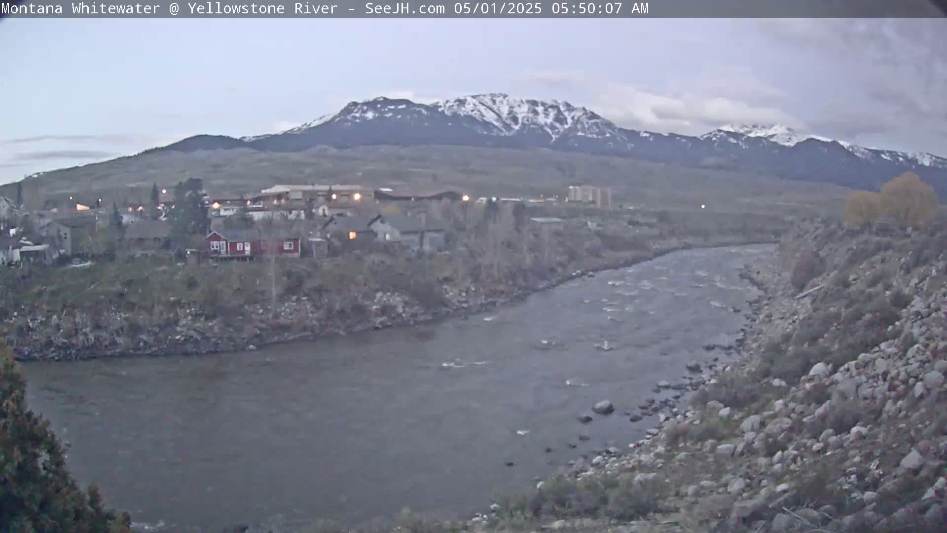 A grayish-blue river flows past a small town nestled beneath snow-capped mountains under an overcast sky.