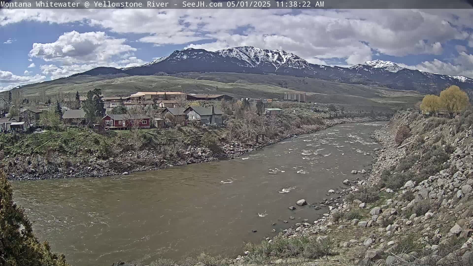 A partly cloudy day reveals a snow-capped mountain range overlooking a river flowing past a small town.