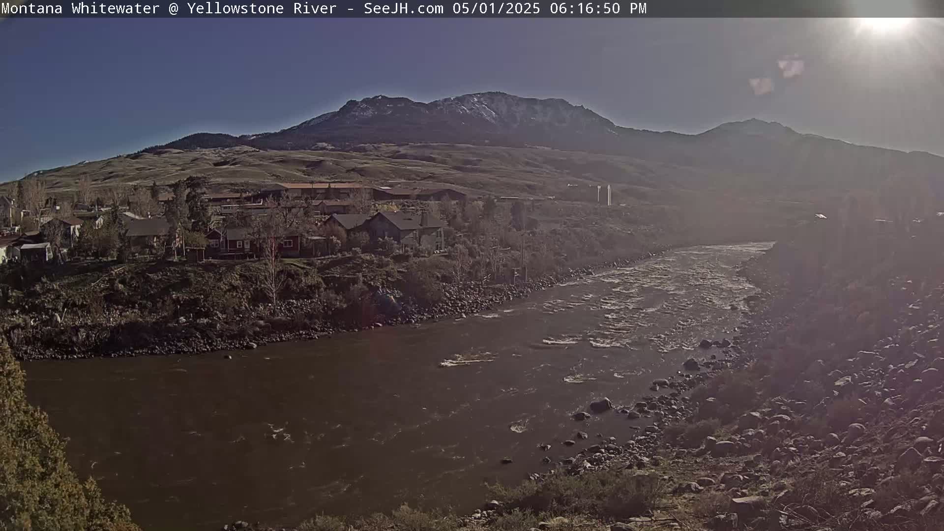 A river flows through a valley under a sunny sky, past a town nestled against snow-capped mountains.