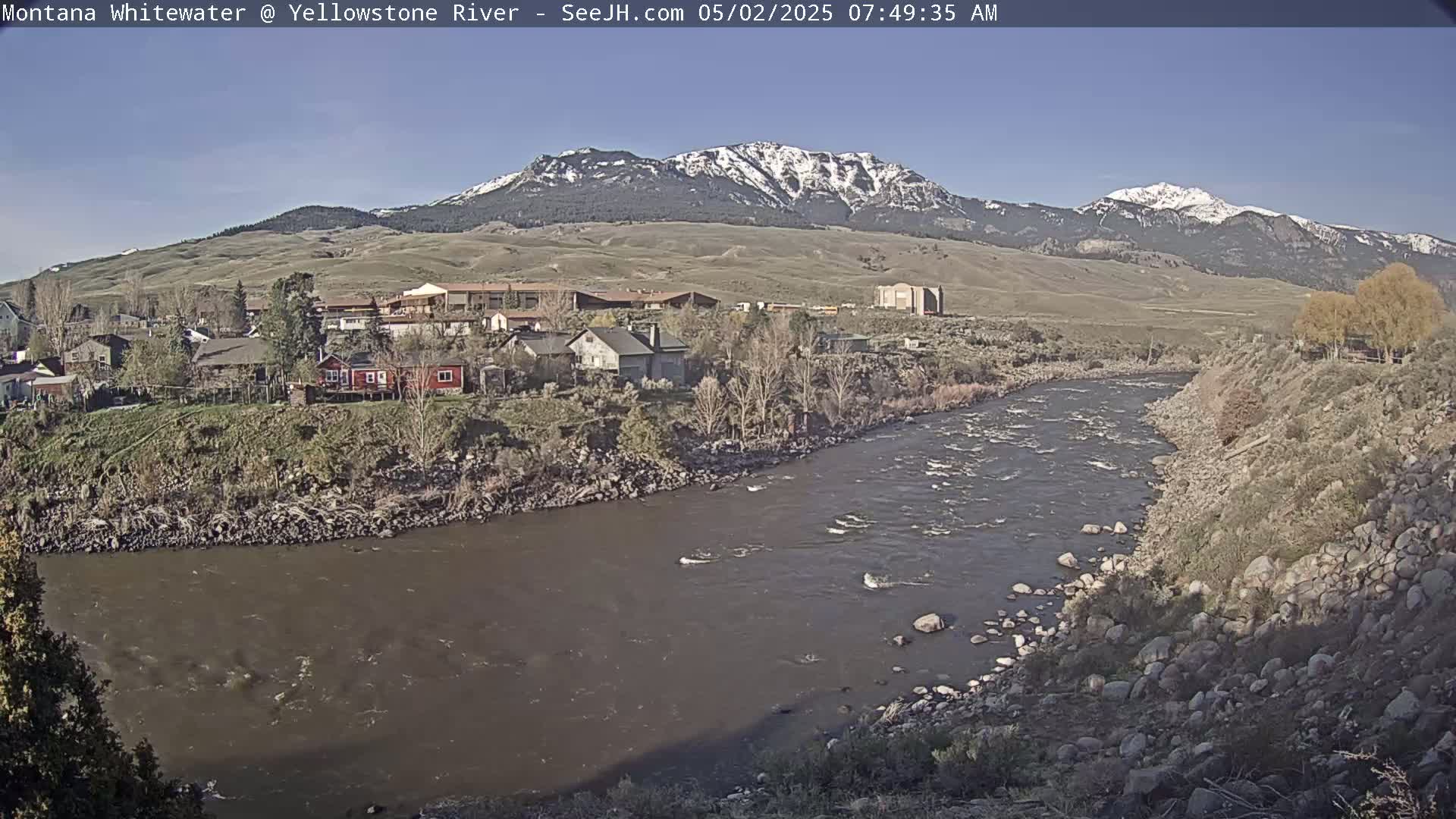 A river flows through a town nestled at the base of snow-capped mountains under a clear sky.