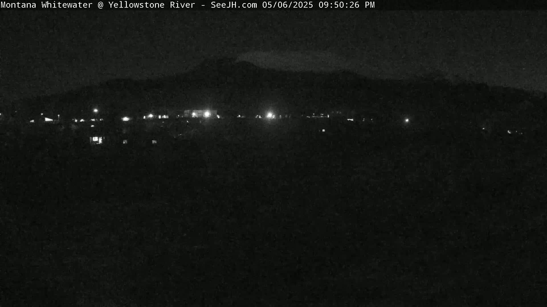 A nighttime, low-light view of a dark landscape with a line of distant lights and a dark mountain in the background.