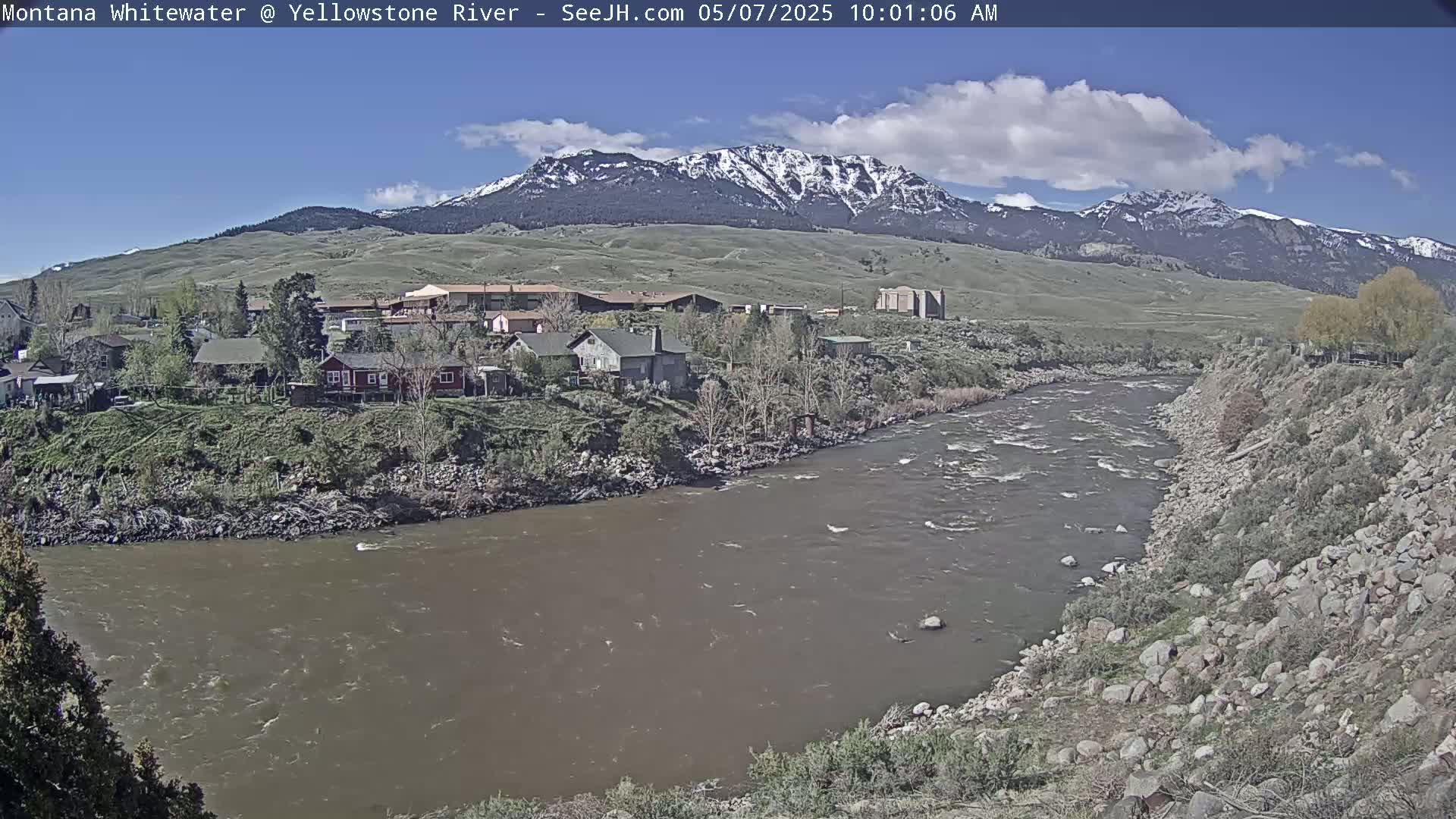 A muddy river flows past a small town nestled at the base of snow-capped mountains under a mostly sunny sky.