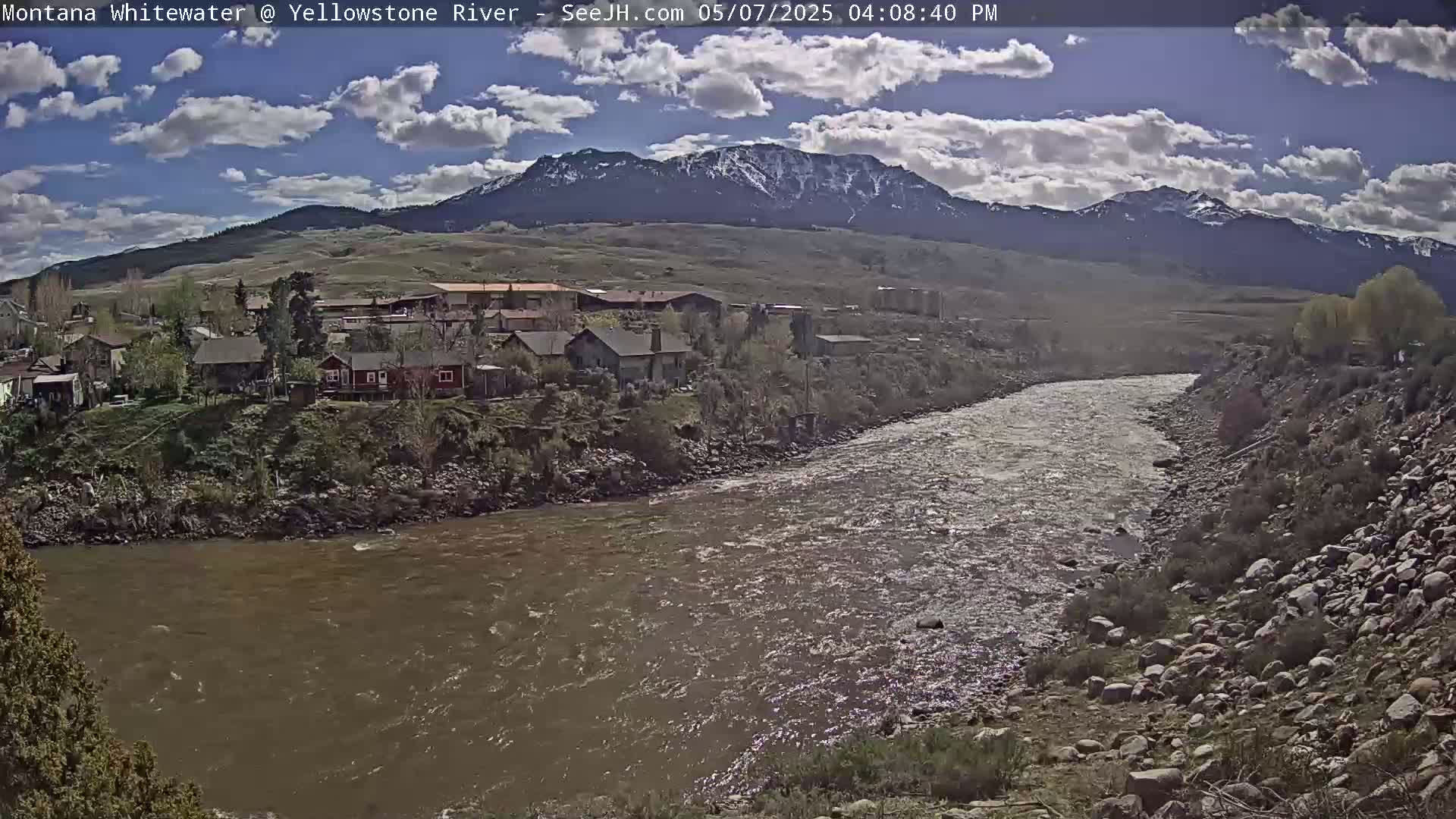 A partly cloudy day shows a wide, brown river flowing past a small town nestled beside snow-capped mountains.