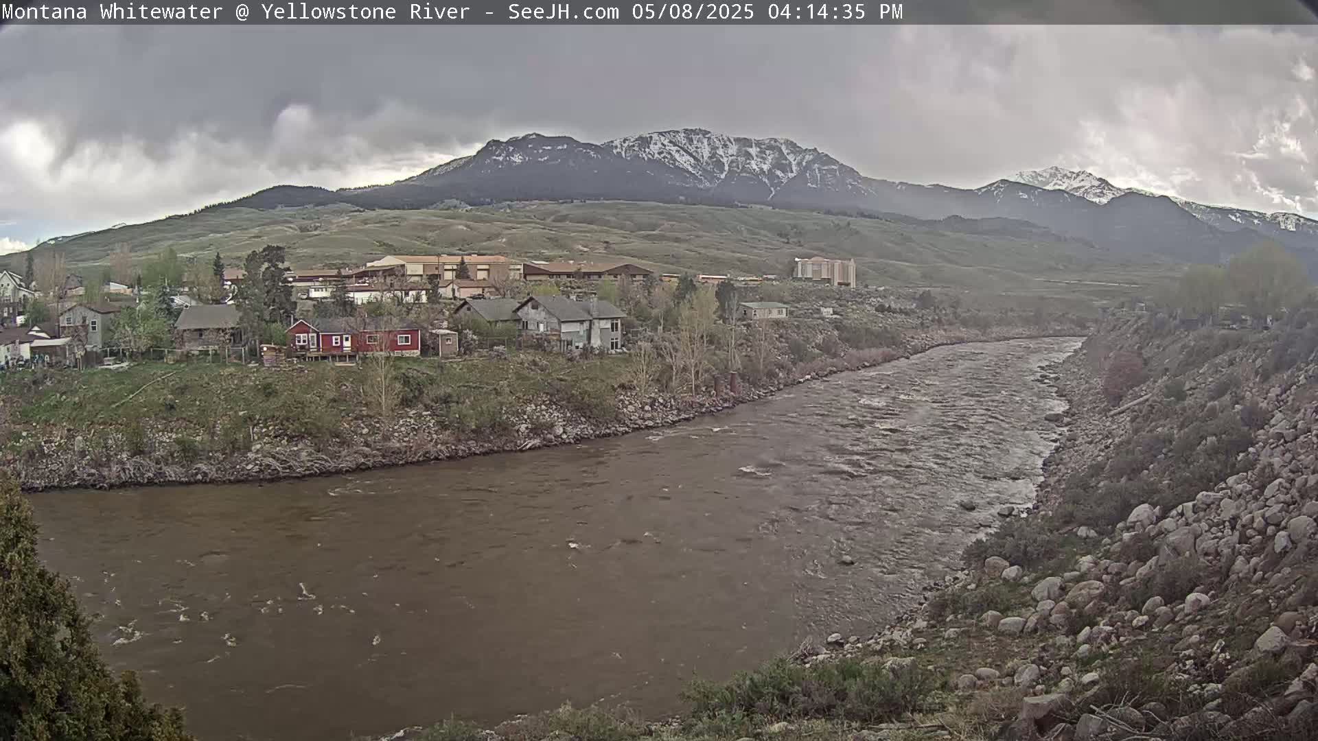 A murky river flows through a town nestled at the base of snow-capped mountains under an overcast sky.