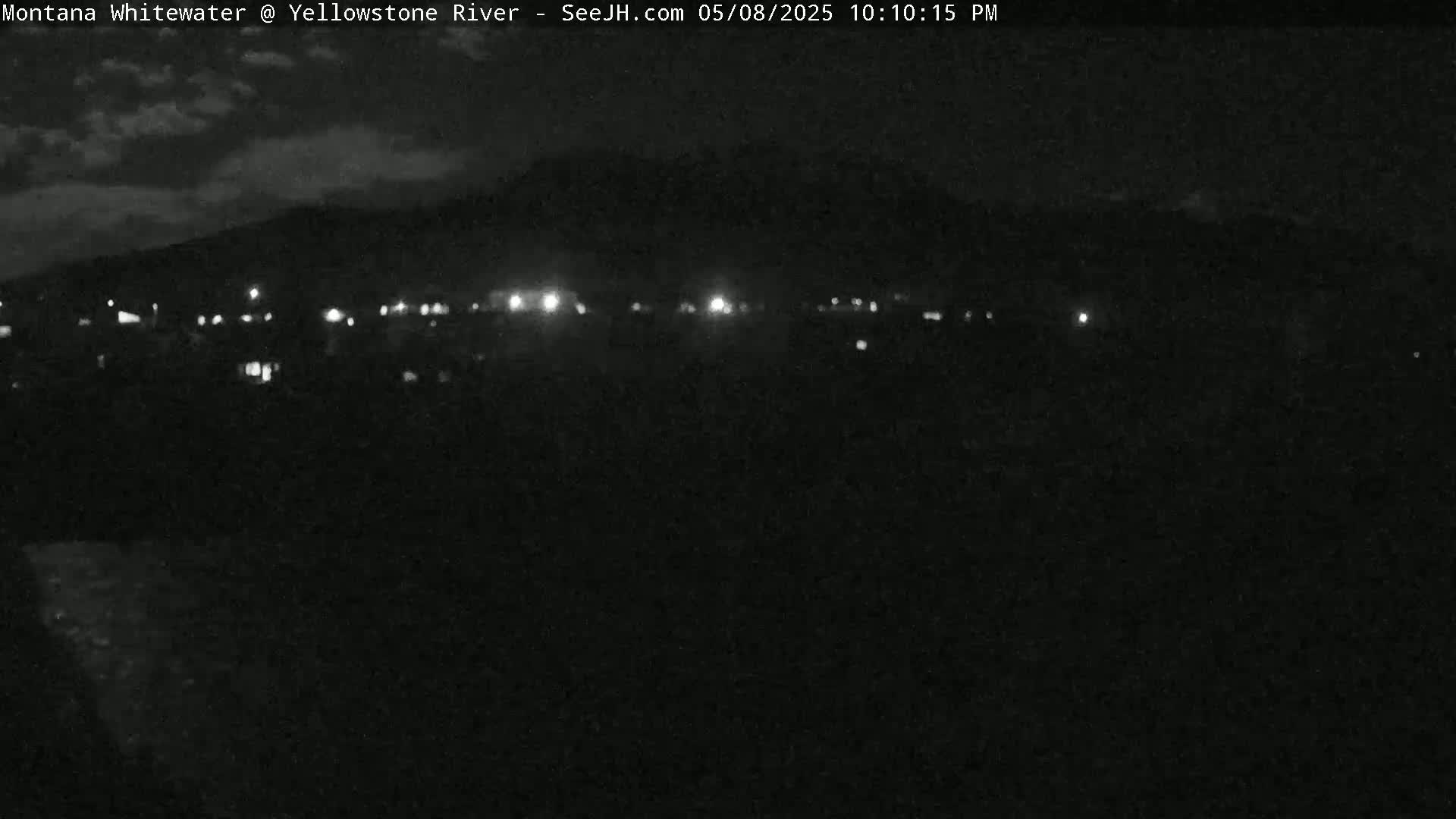 A nighttime, low-light view shows a distant line of lights against a dark, cloudy sky and silhouetted hills.