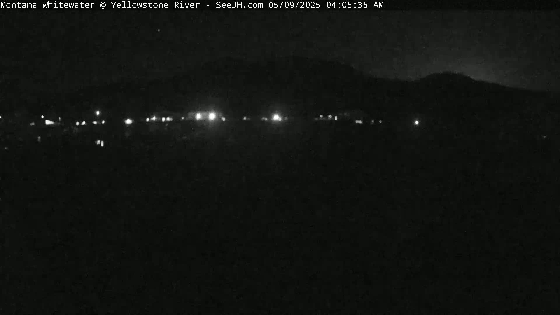 A nighttime view shows a dark landscape with a distant line of lights and a dark hill or mountain in the background under a clear, dark sky.