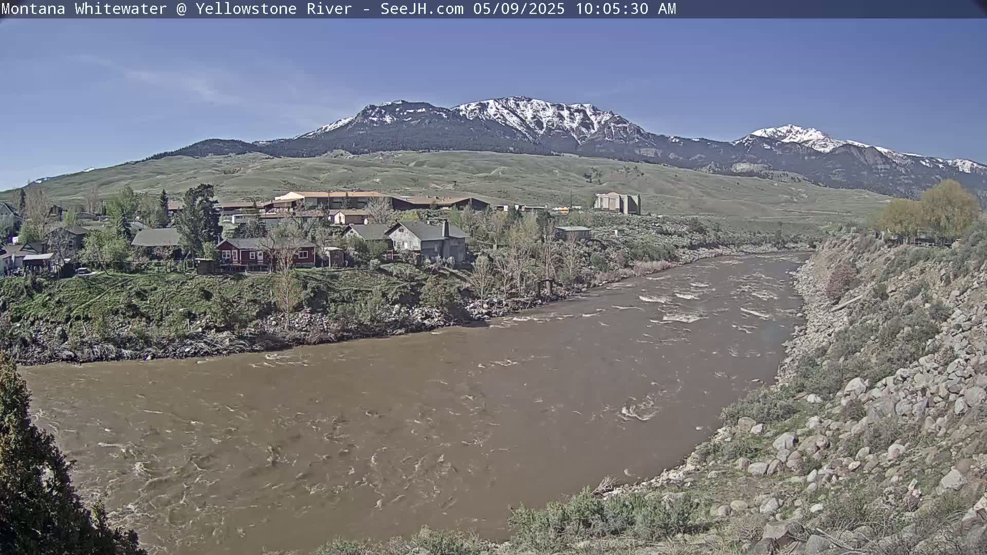 A muddy river flows past a small town nestled at the base of snow-capped mountains under a clear sky.