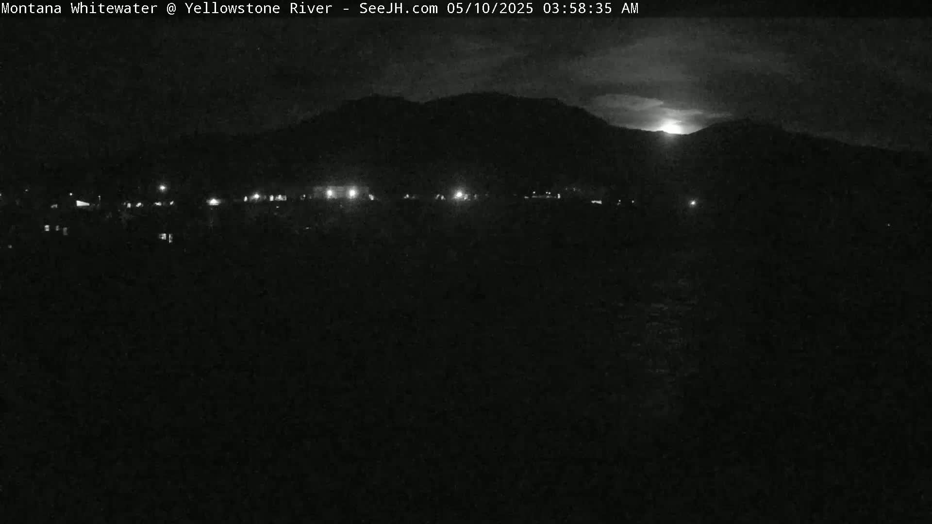 A nighttime, low-light view shows a dark landscape with a distant, silhouetted mountain range under a partly cloudy sky, with a few faint lights visible in the foreground.