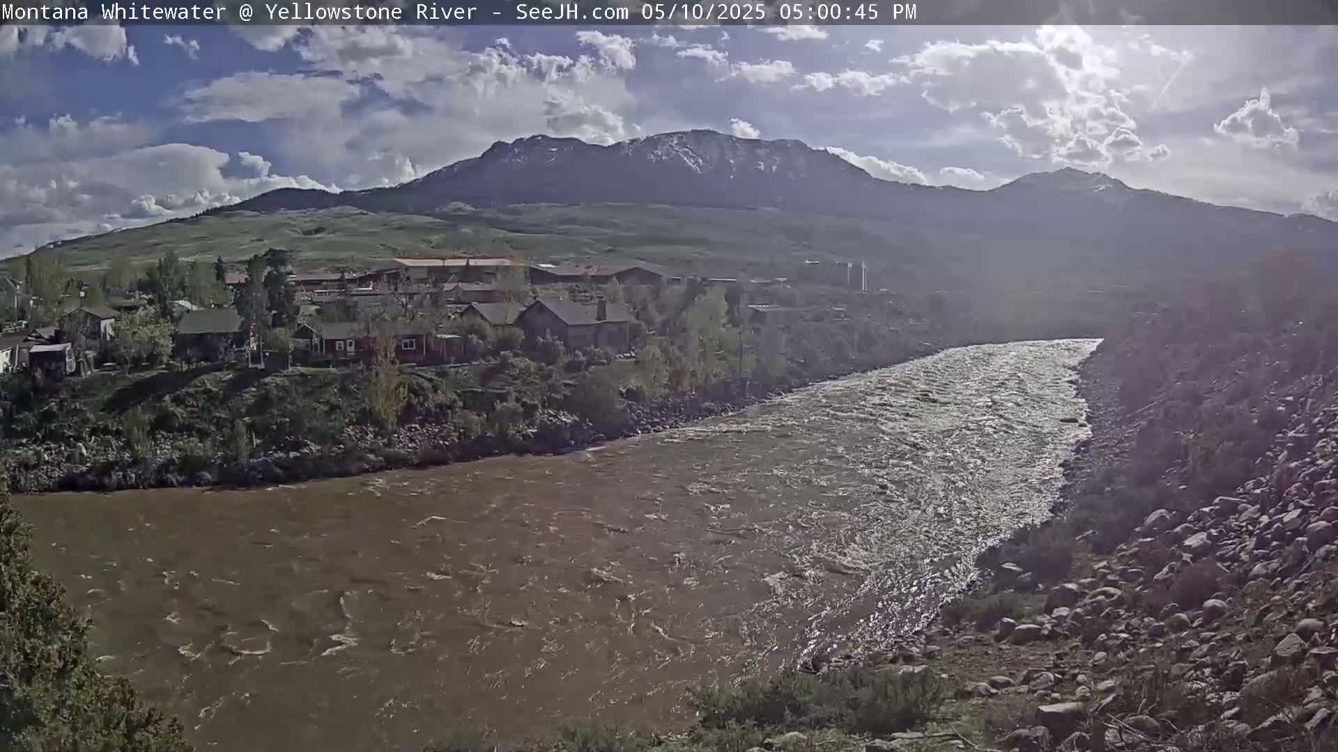 A muddy river flows past a small town nestled beneath a mountain range under a partly cloudy sky.