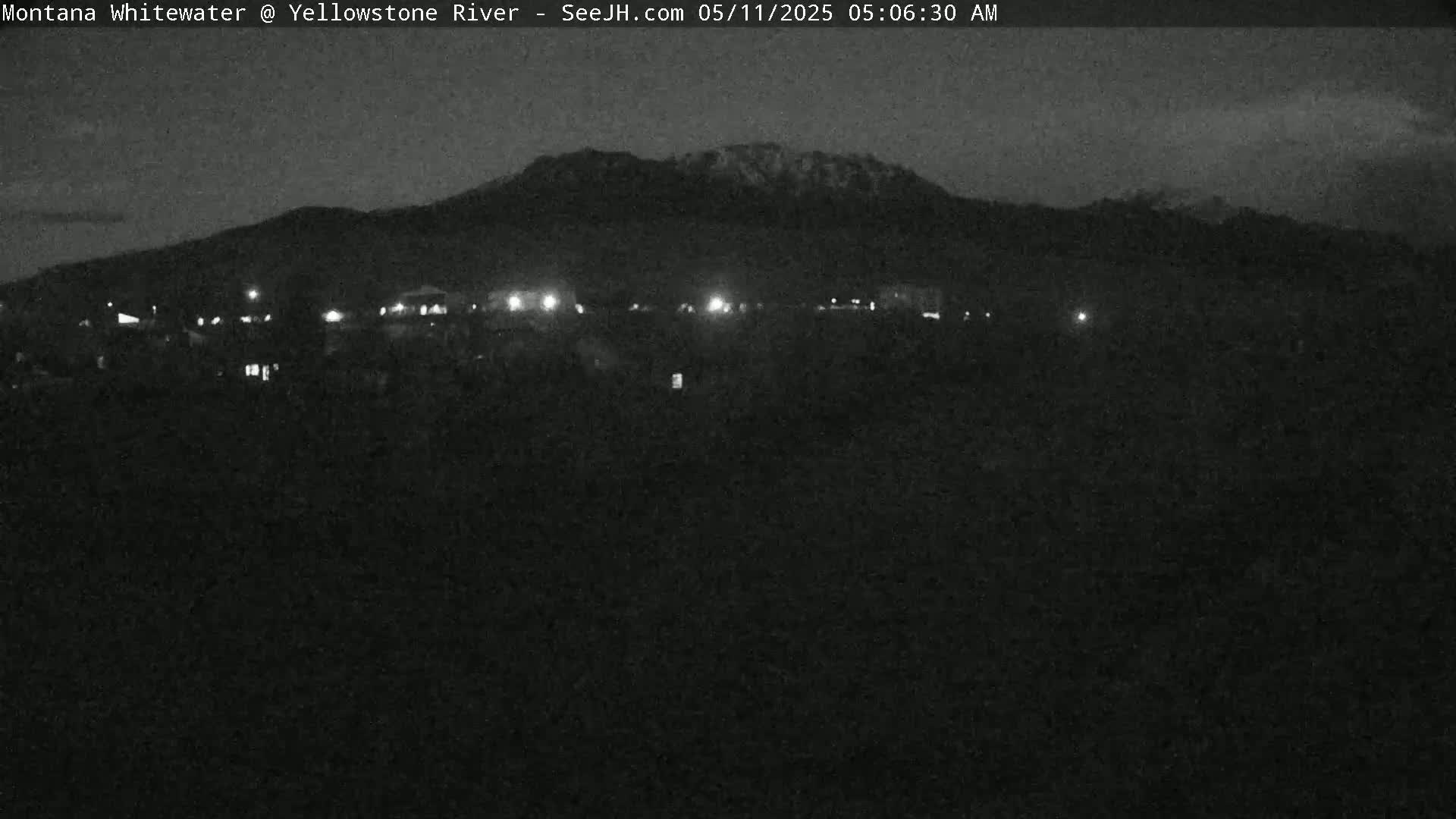 A nighttime, low-light view of a snow-capped mountain range behind a dark, flat landscape with sparsely lit buildings in the foreground.