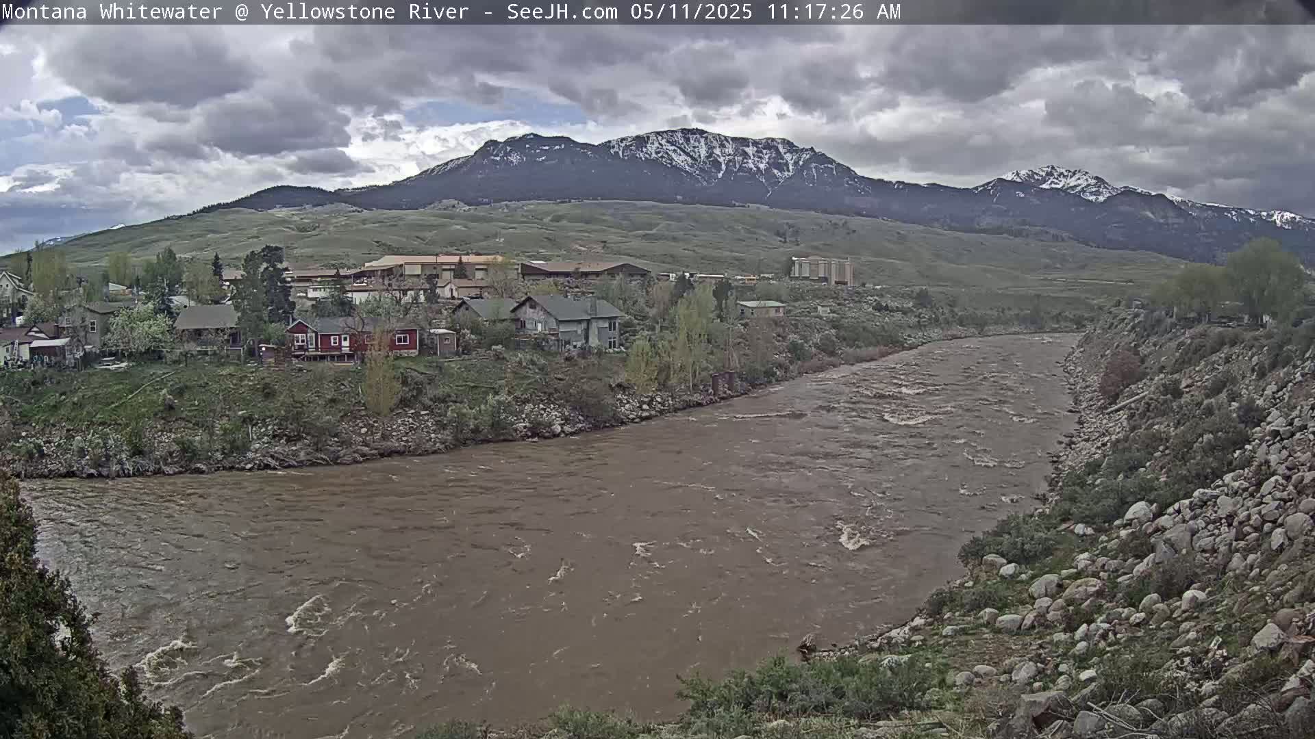 A muddy river flows past a small town nestled at the base of a snow-capped mountain range under a cloudy sky.