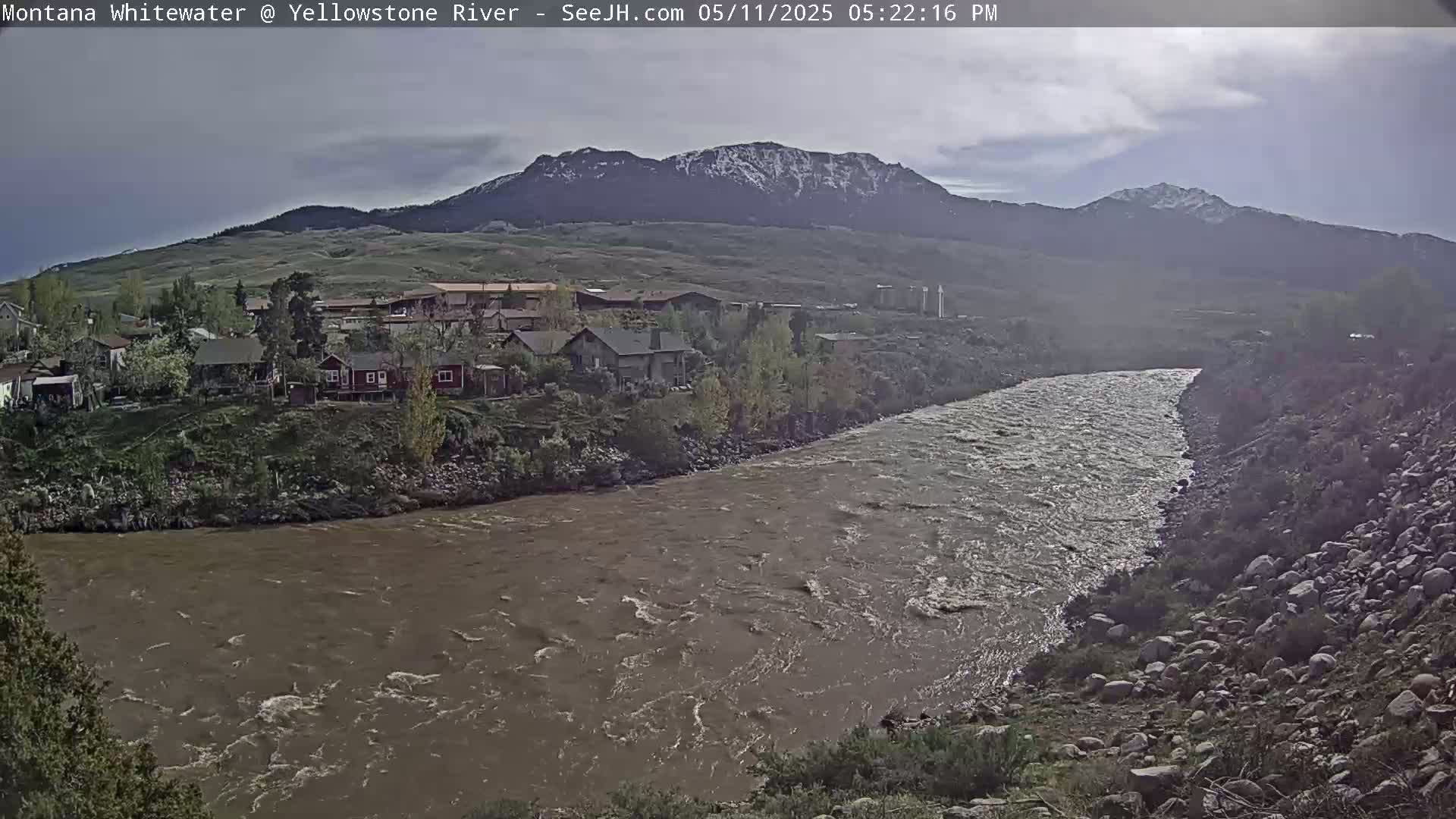 A muddy river flows past a residential area nestled against snow-capped mountains under a cloudy sky.