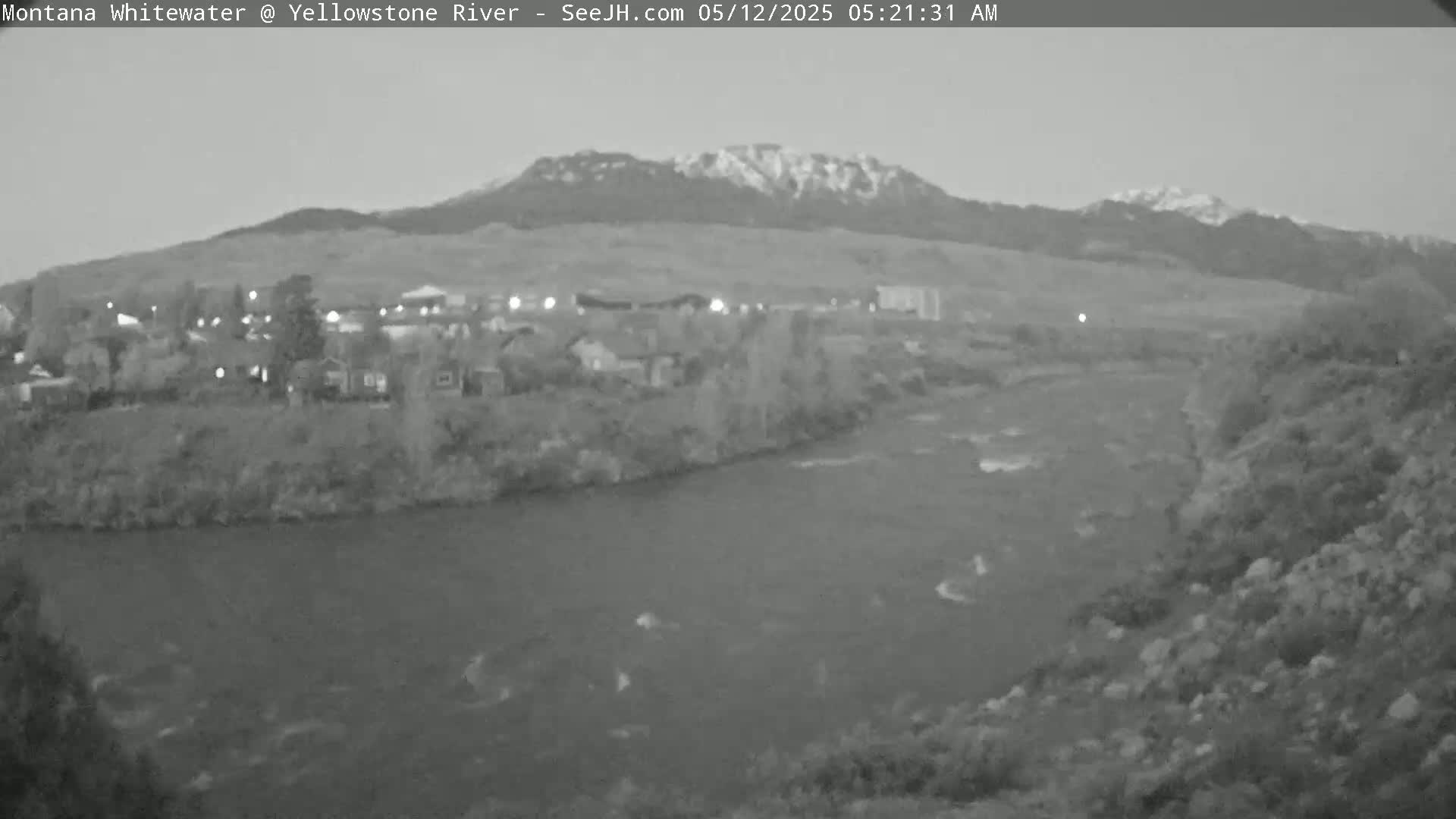 A grayscale image shows a river flowing past a town nestled at the base of snow-capped mountains under a dawn sky.