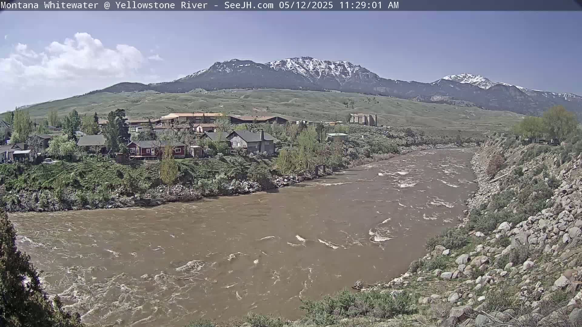 A muddy river flows past a small town nestled at the base of snow-capped mountains under a mostly clear sky.