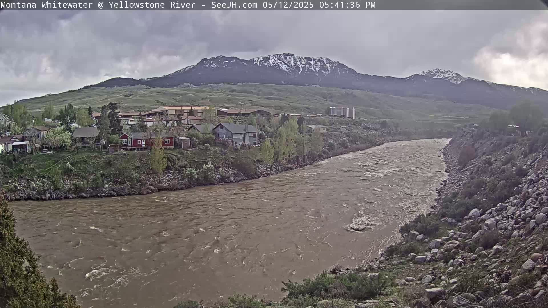 A muddy, swiftly flowing river curves past a small town nestled at the base of snow-capped mountains under a cloudy sky.