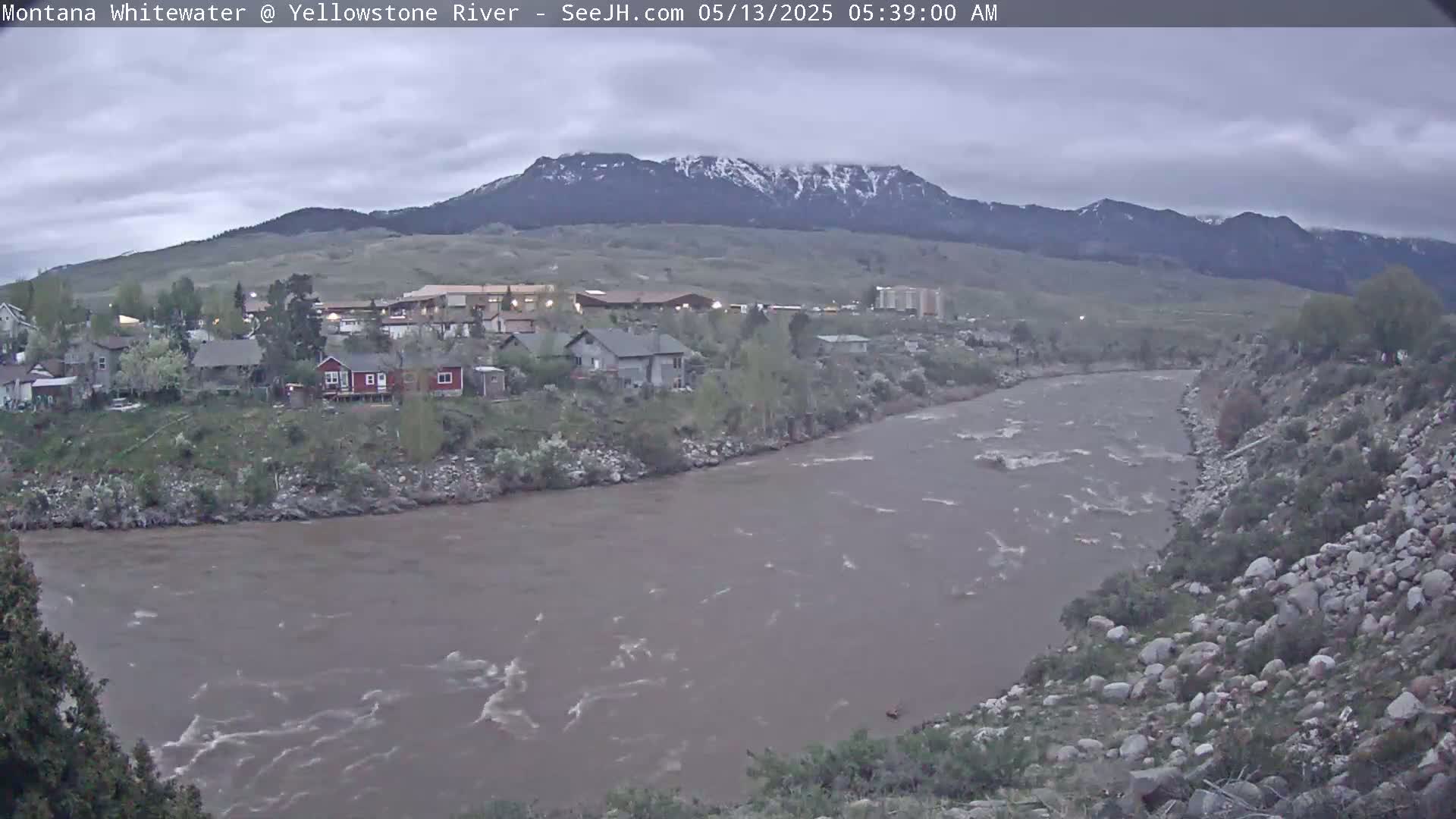 A wide, muddy river flows past a small town nestled against a mountain range with some snow on the peaks under an overcast sky.