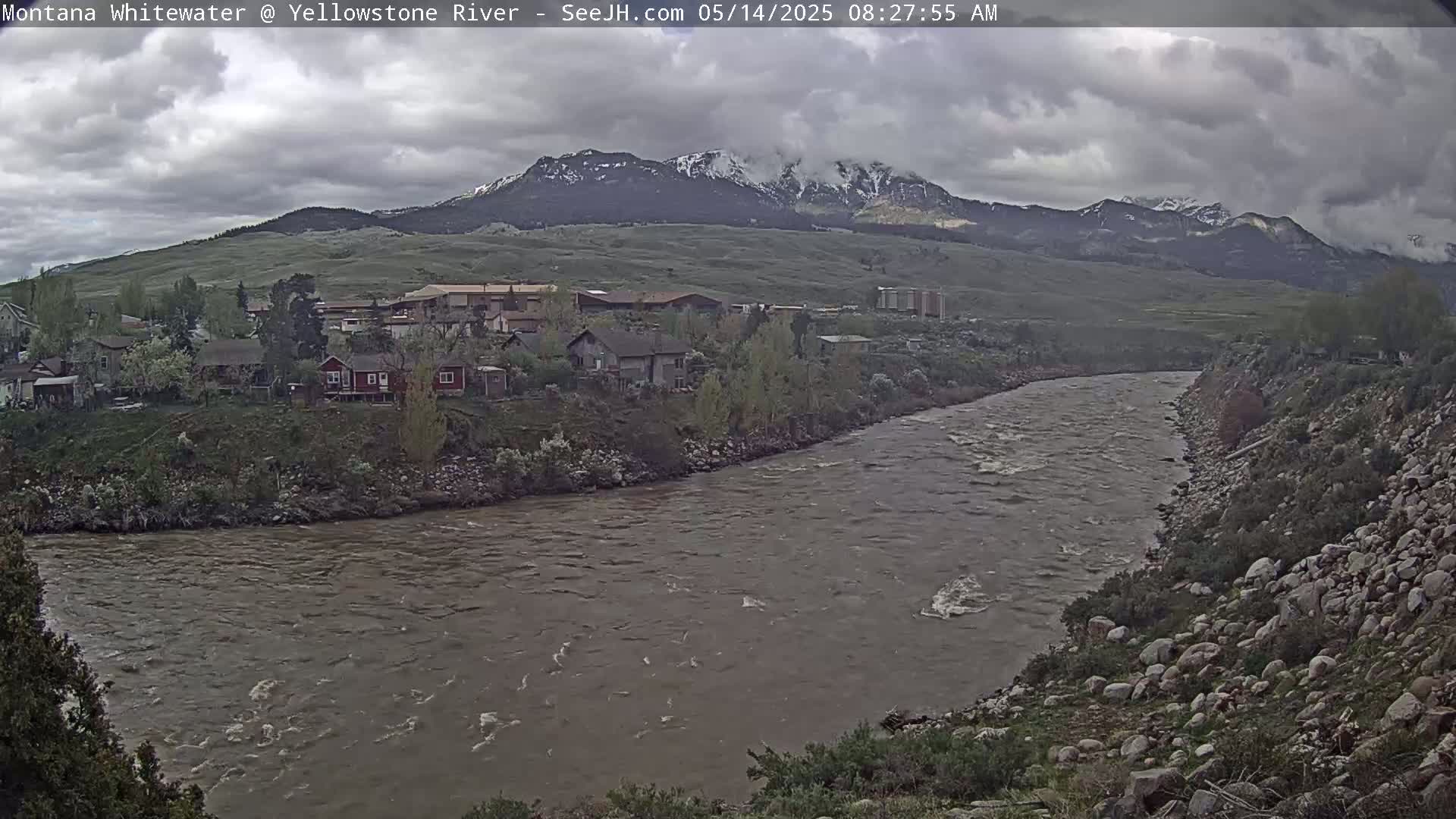 A wide, muddy river flows past a small town nestled at the base of snow-capped mountains under a cloudy sky.