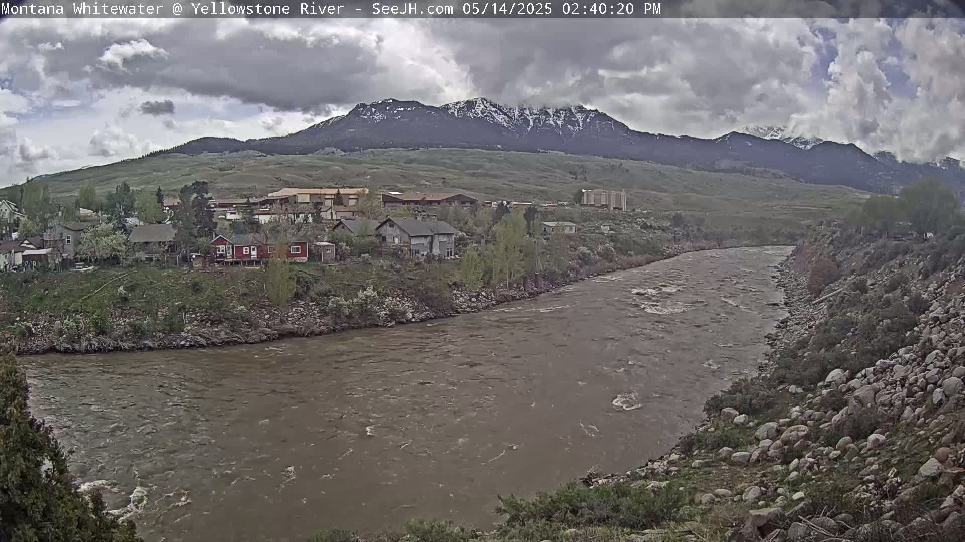 A wide, muddy river flows past a small town nestled at the base of snow-capped mountains under a cloudy sky.