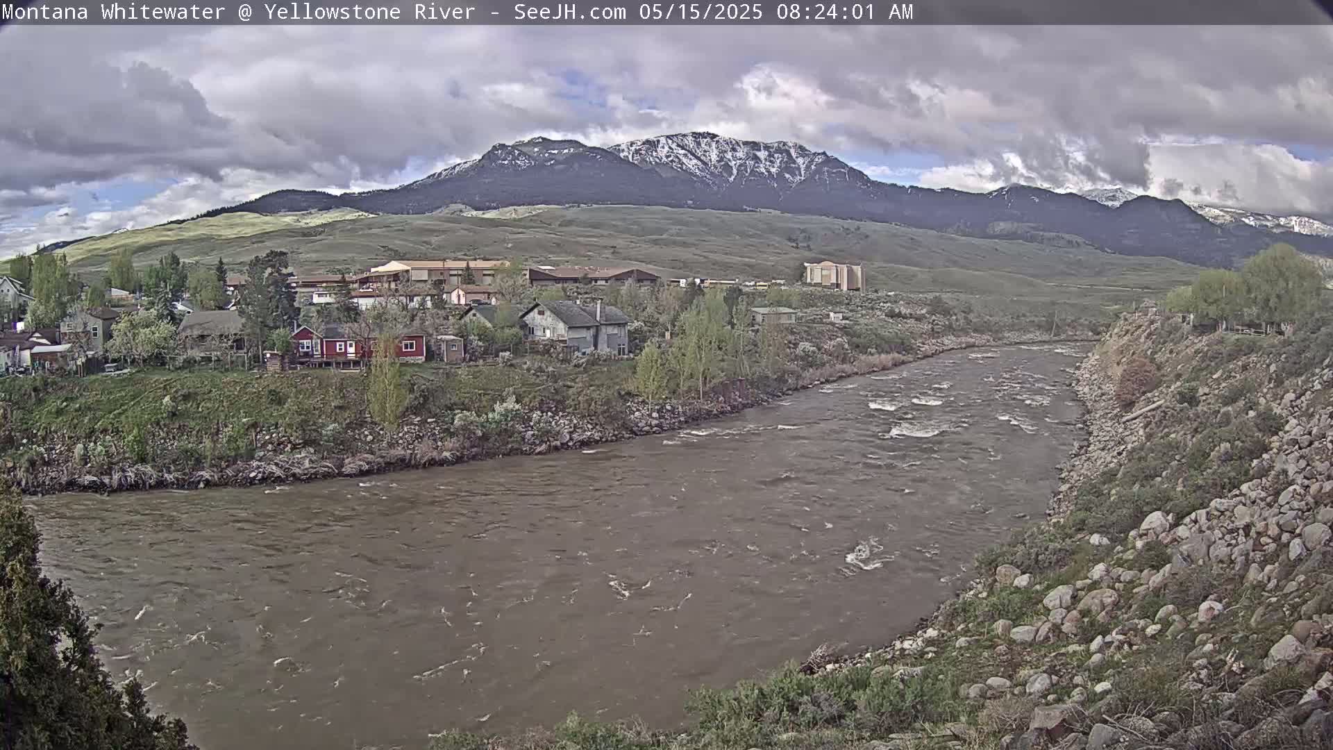 A cloudy day shows a swiftly flowing river curving past a small town nestled against a mountain range with snow-capped peaks.