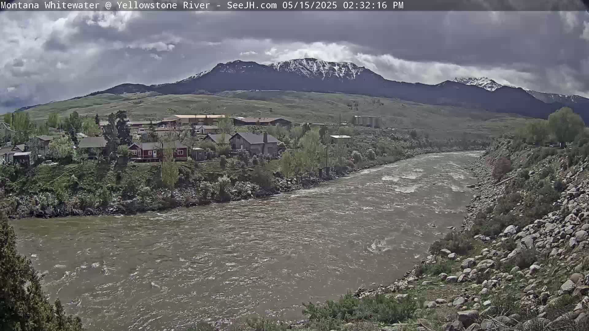 A swiftly flowing river curves past a rocky bank and houses nestled amongst greenery, with a cloudy sky and snow-capped mountains in the background.
