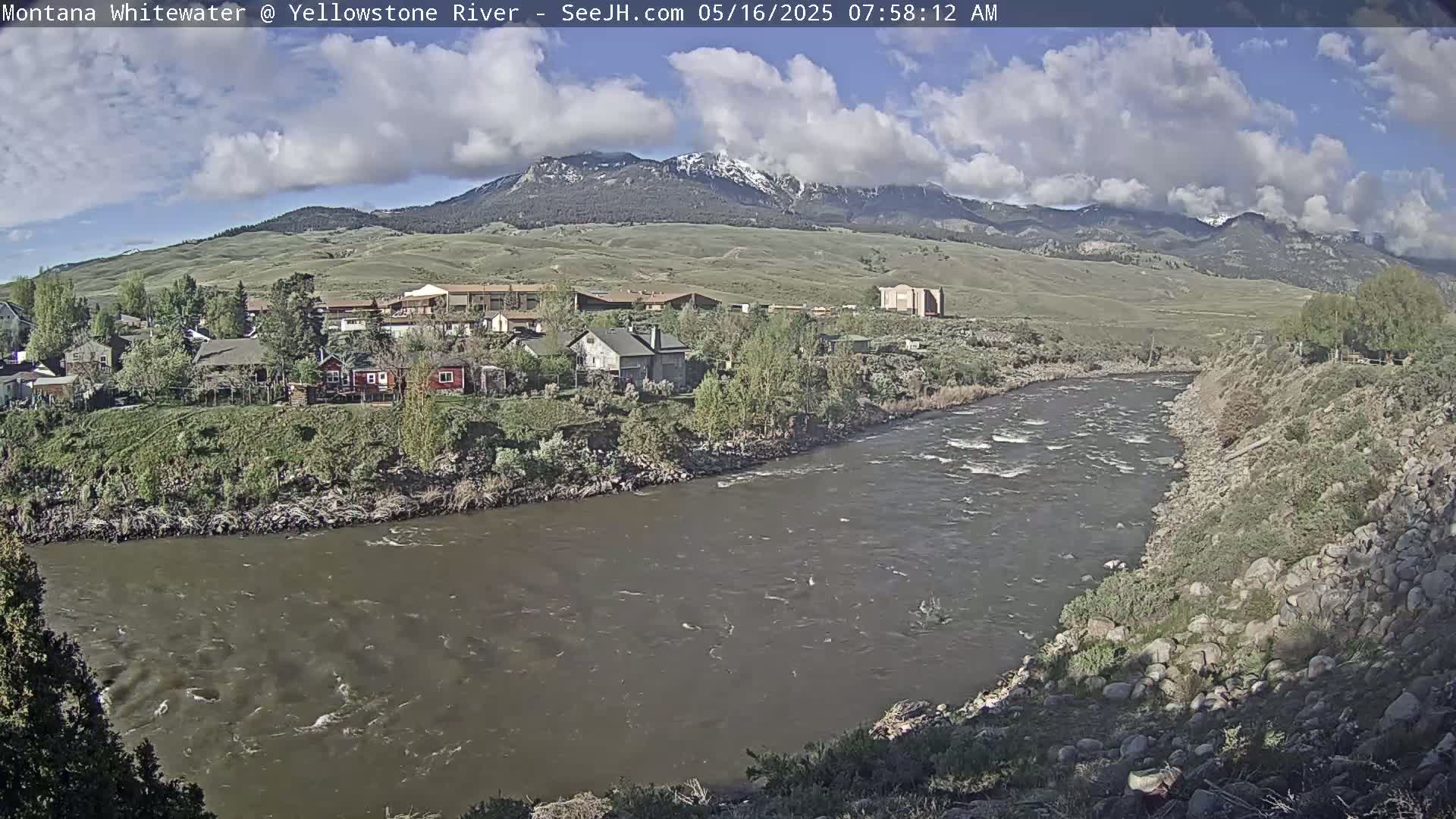 A partly cloudy day shows a river flowing past a small town nestled at the base of a snow-capped mountain range.