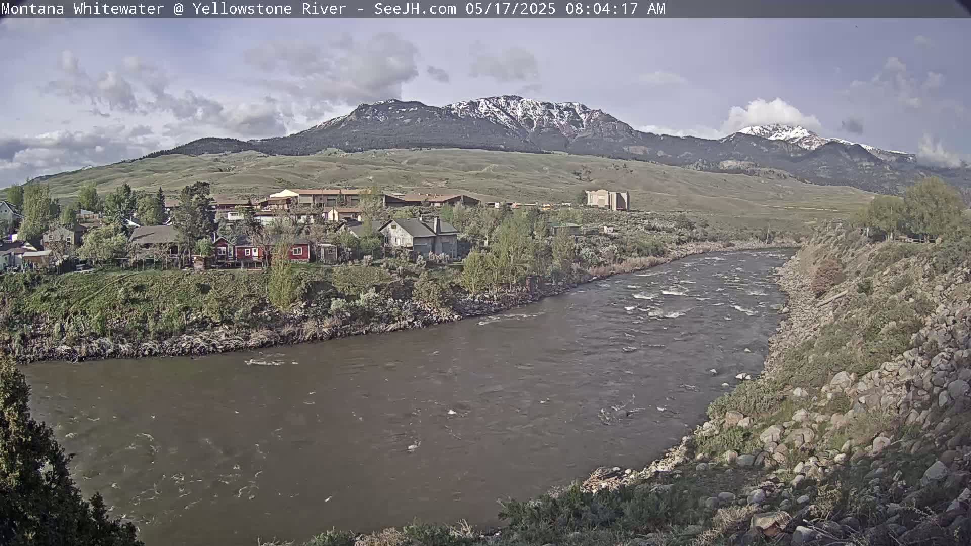 A partly cloudy day reveals a river flowing past a small town nestled against snow-capped mountains.