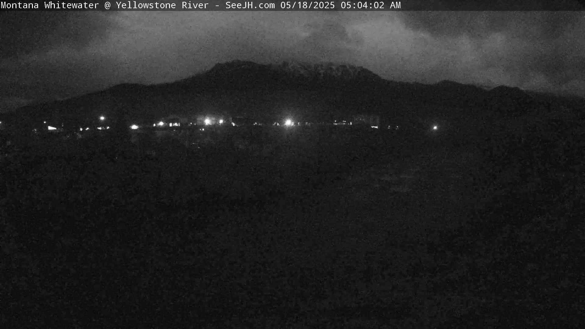 A nighttime, overcast view shows a dark, flat landscape with a line of distant lights and a mountain range in the background.