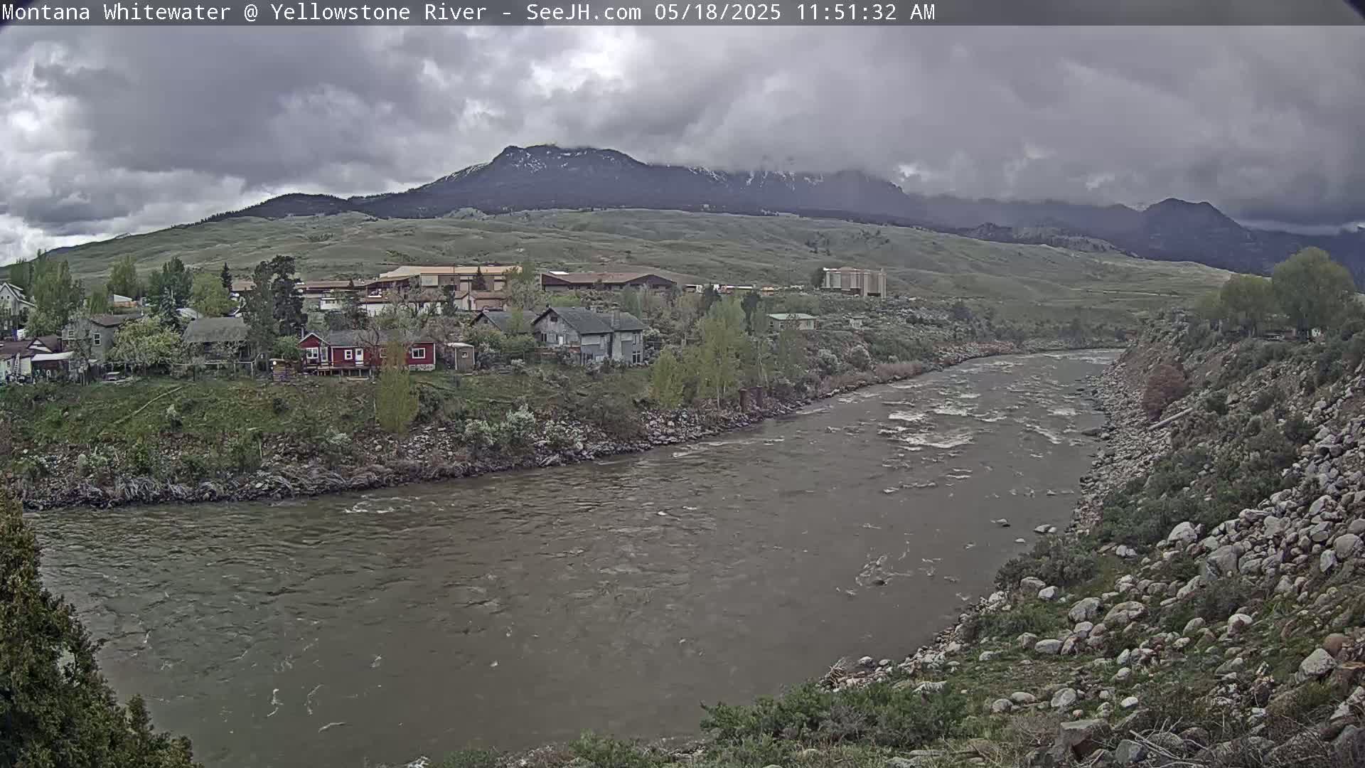 A river flows through a town nestled at the base of a mountain range under a cloudy sky.