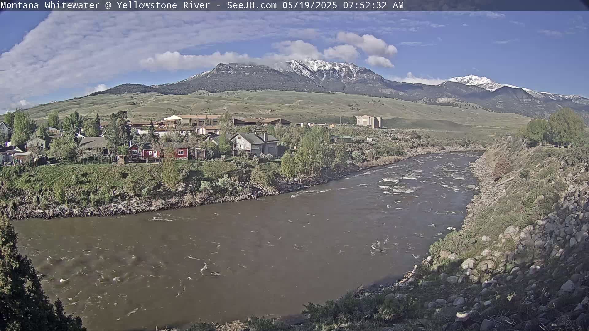 A partly cloudy day shows a river flowing past a small town nestled against snow-capped mountains.