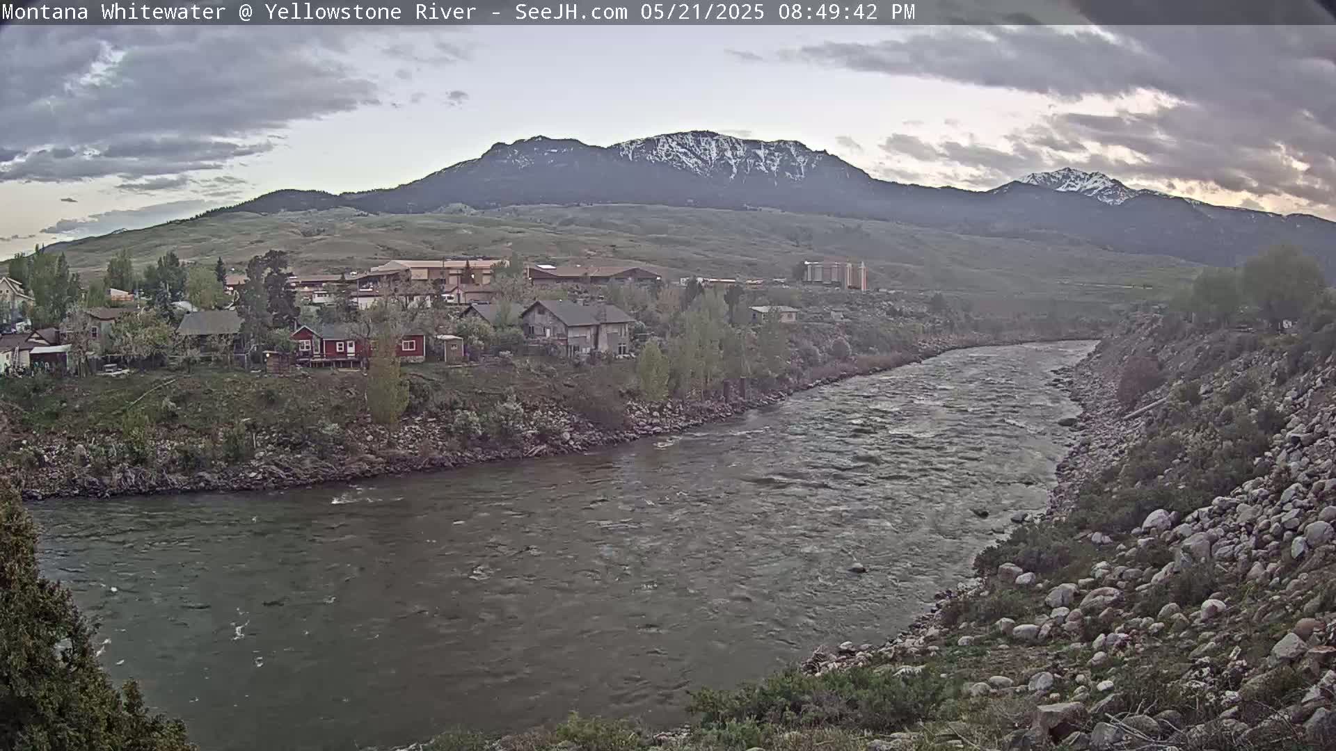 A wide, flowing river curves past a small town nestled at the base of a snow-capped mountain range under a partly cloudy sky.