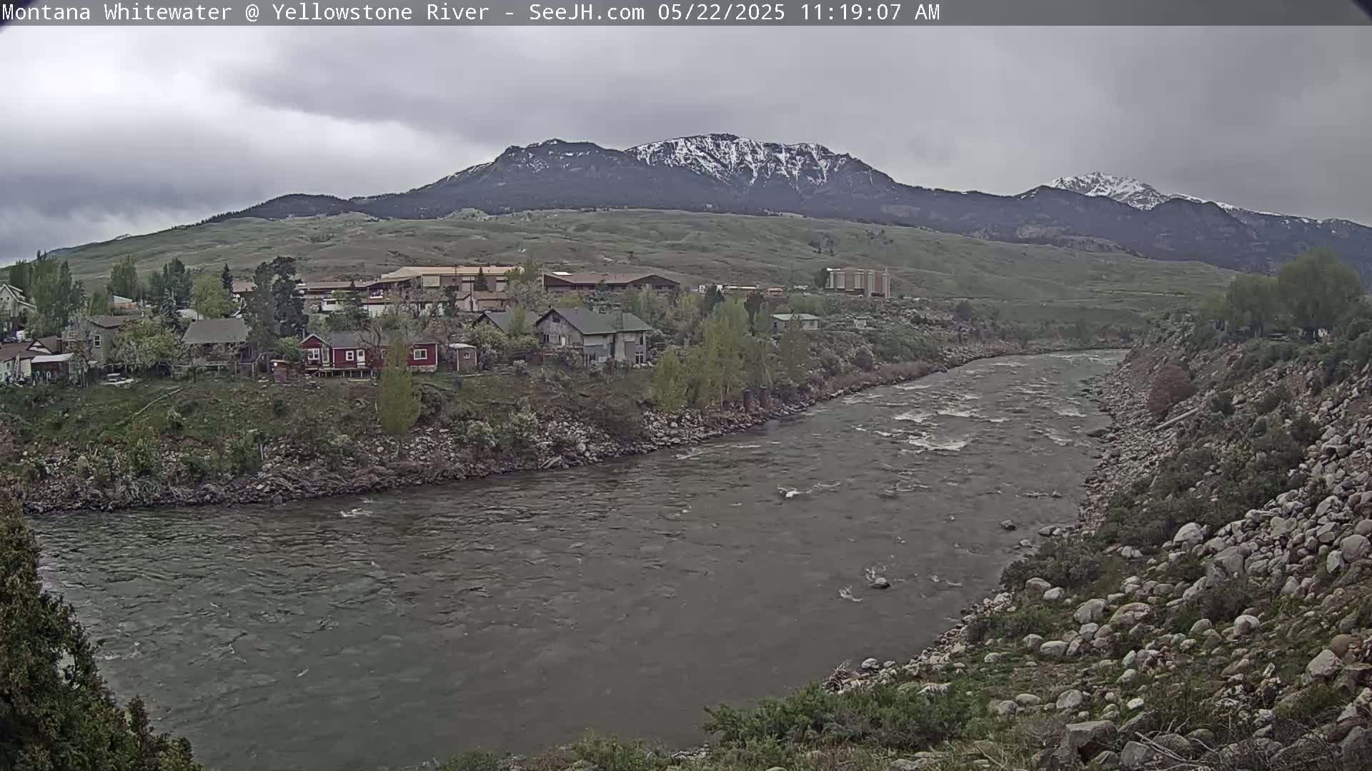 A grayish river flows past a small town nestled at the base of a snow-capped mountain range under an overcast sky.