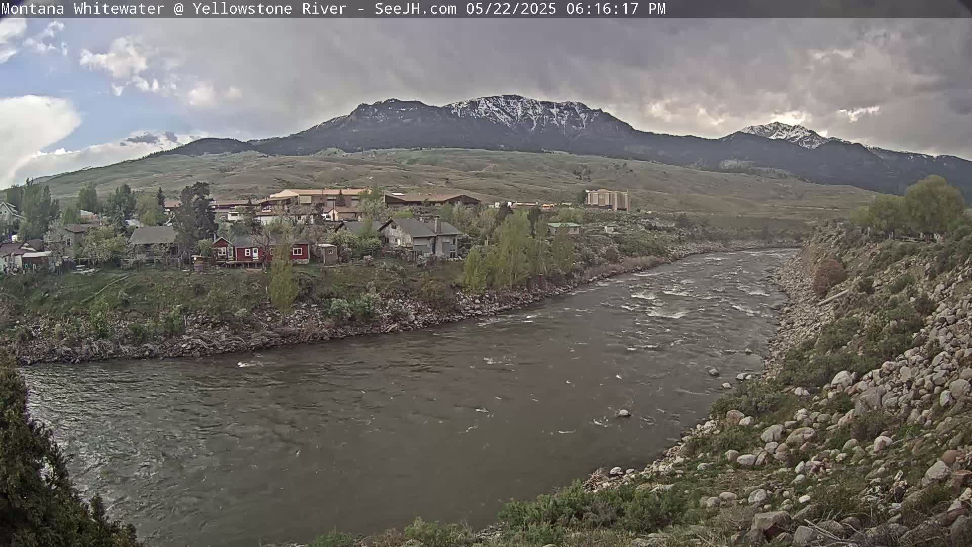 A river flows past a town nestled at the base of snow-capped mountains under a cloudy sky.