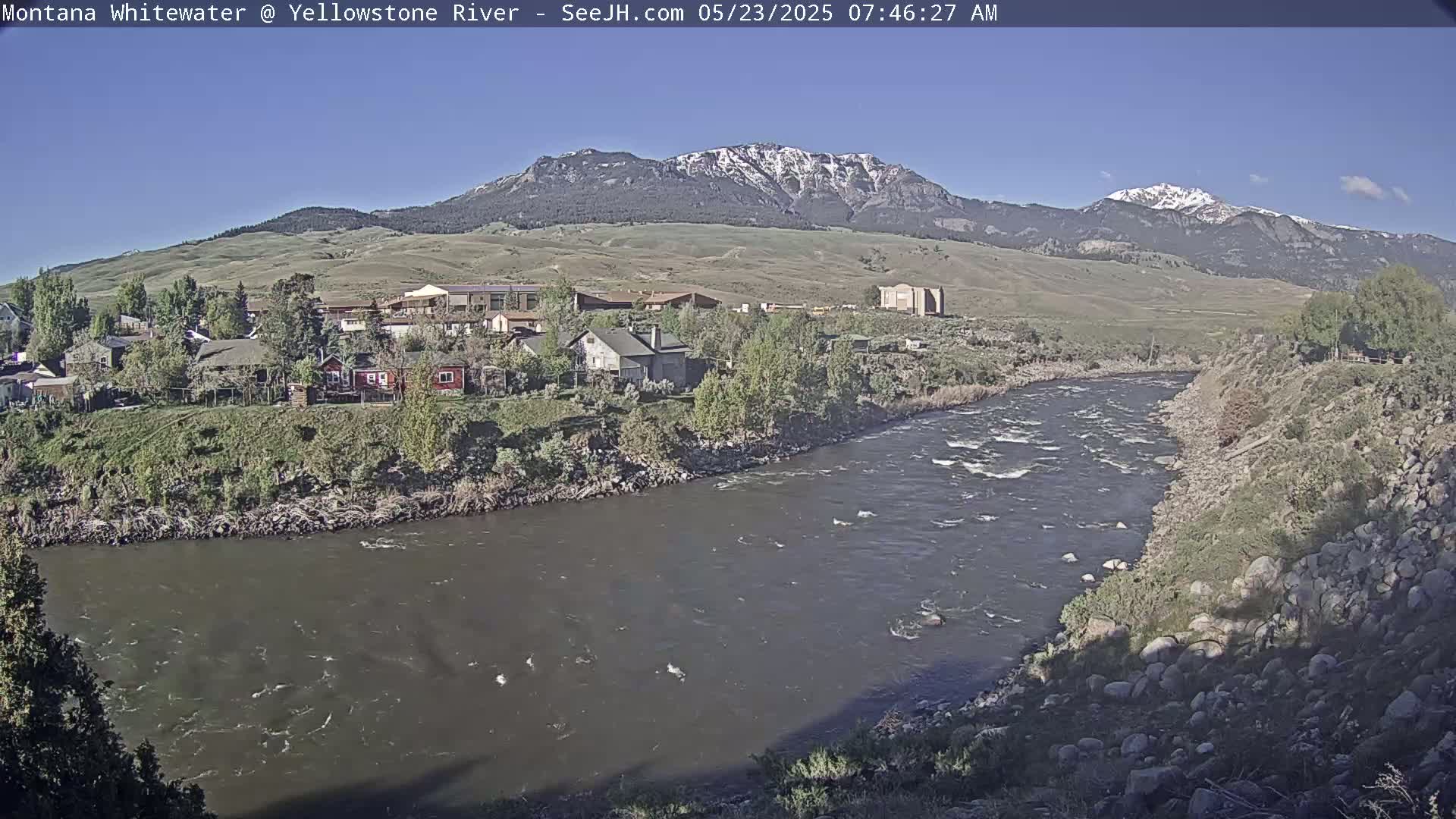 A swiftly flowing river curves past a small town nestled at the base of snow-capped mountains under a clear blue sky.