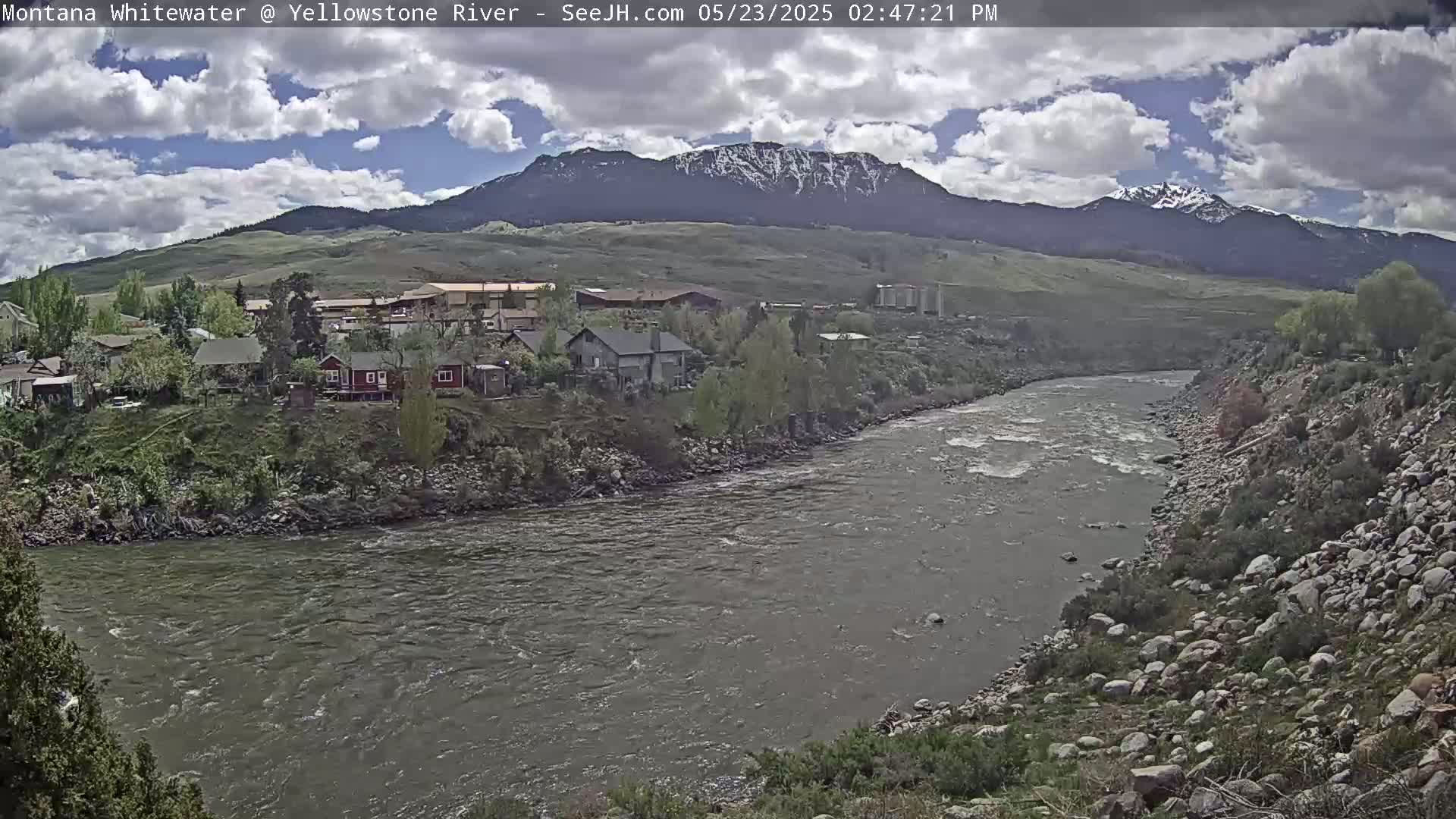 A partly cloudy day shows a river flowing through a valley with houses and a snow-capped mountain range in the background.