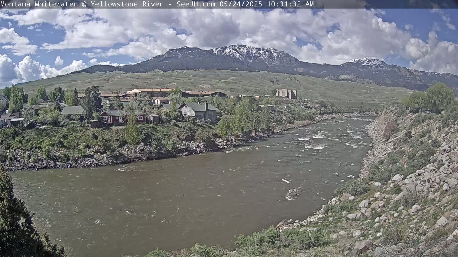 A partly cloudy day reveals a river flowing past a small town nestled at the foot of snow-capped mountains.