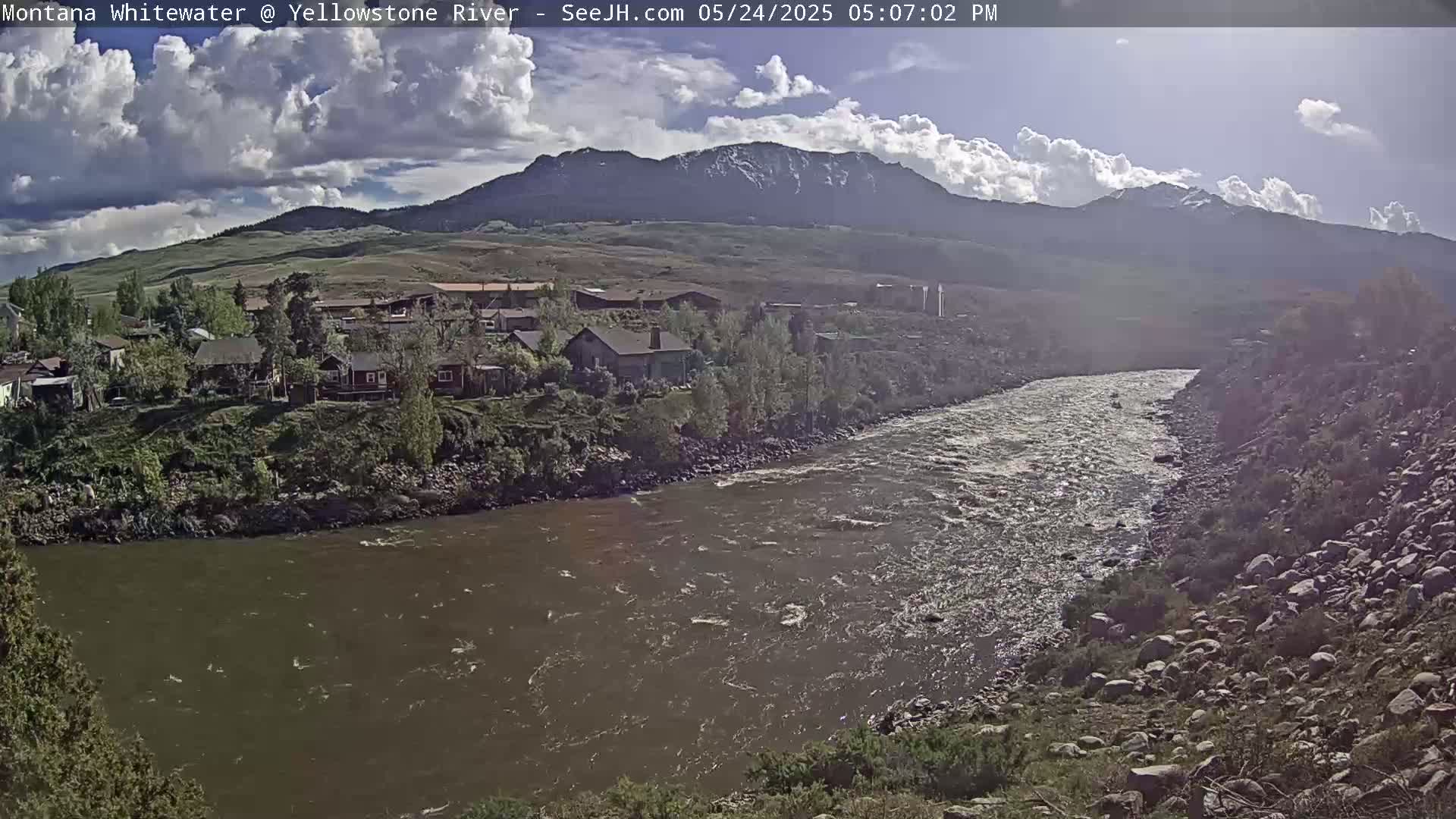 A partly cloudy day reveals a flowing river next to a small town nestled beneath a mountain range with snow-capped peaks.