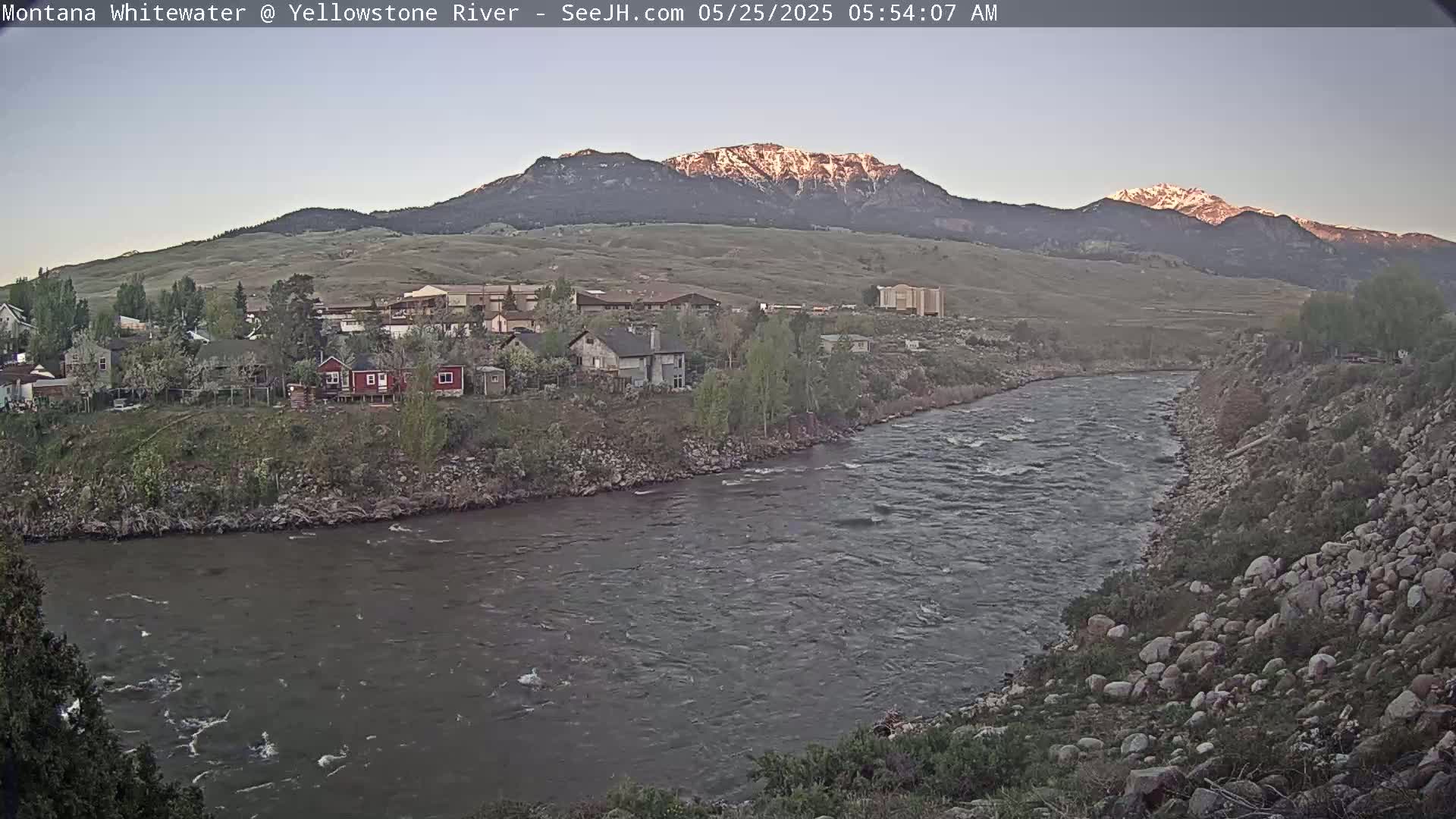 A river flows past houses and a rocky bank, with snow-capped mountains in the background under a clear, twilight sky.